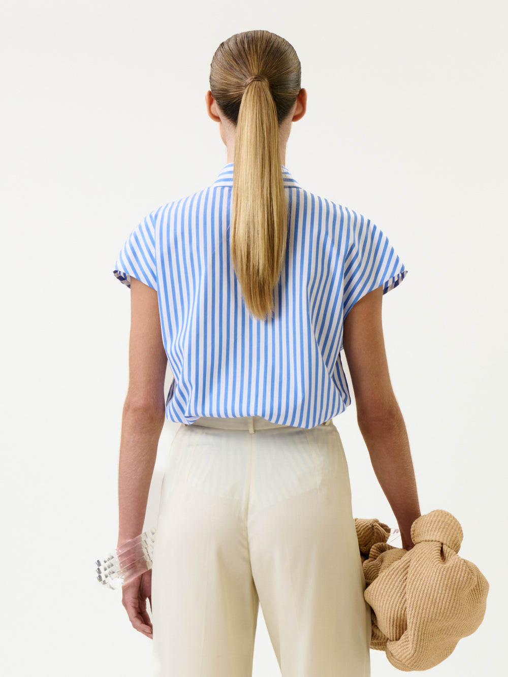 A woman with a sleek ponytail is wearing the Maribel Shirt and cream pants. She is facing away and holding a textured beige object in her left hand, against a plain light background.
