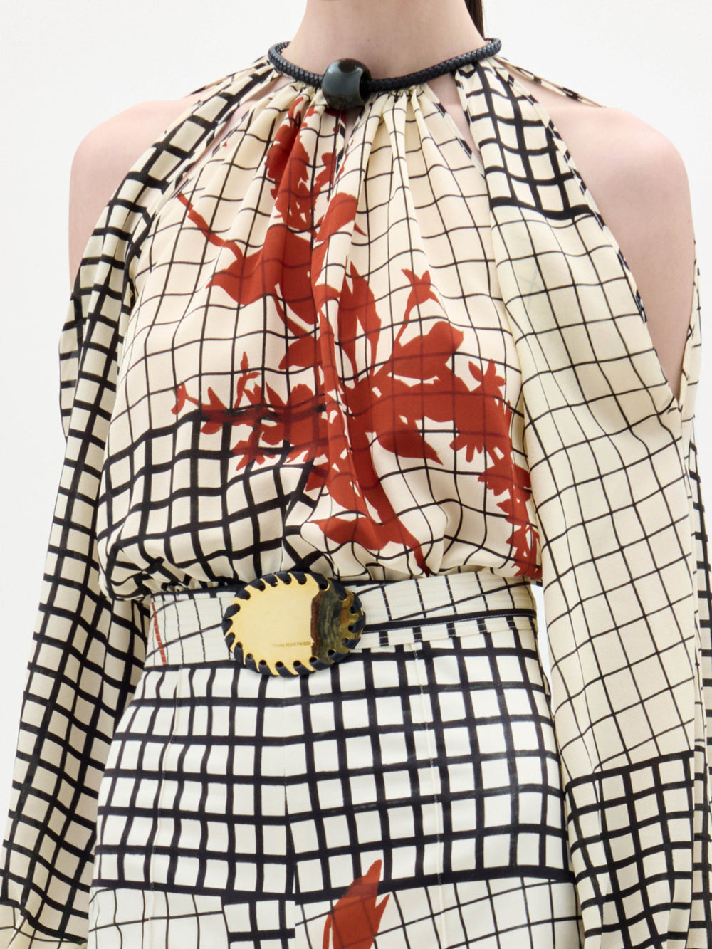 A woman wears the Elke Blouse, a cream and black grid-patterned top with red floral accents, cold-shoulder sleeves, and a belt with a circular gold buckle, standing against a plain white background.