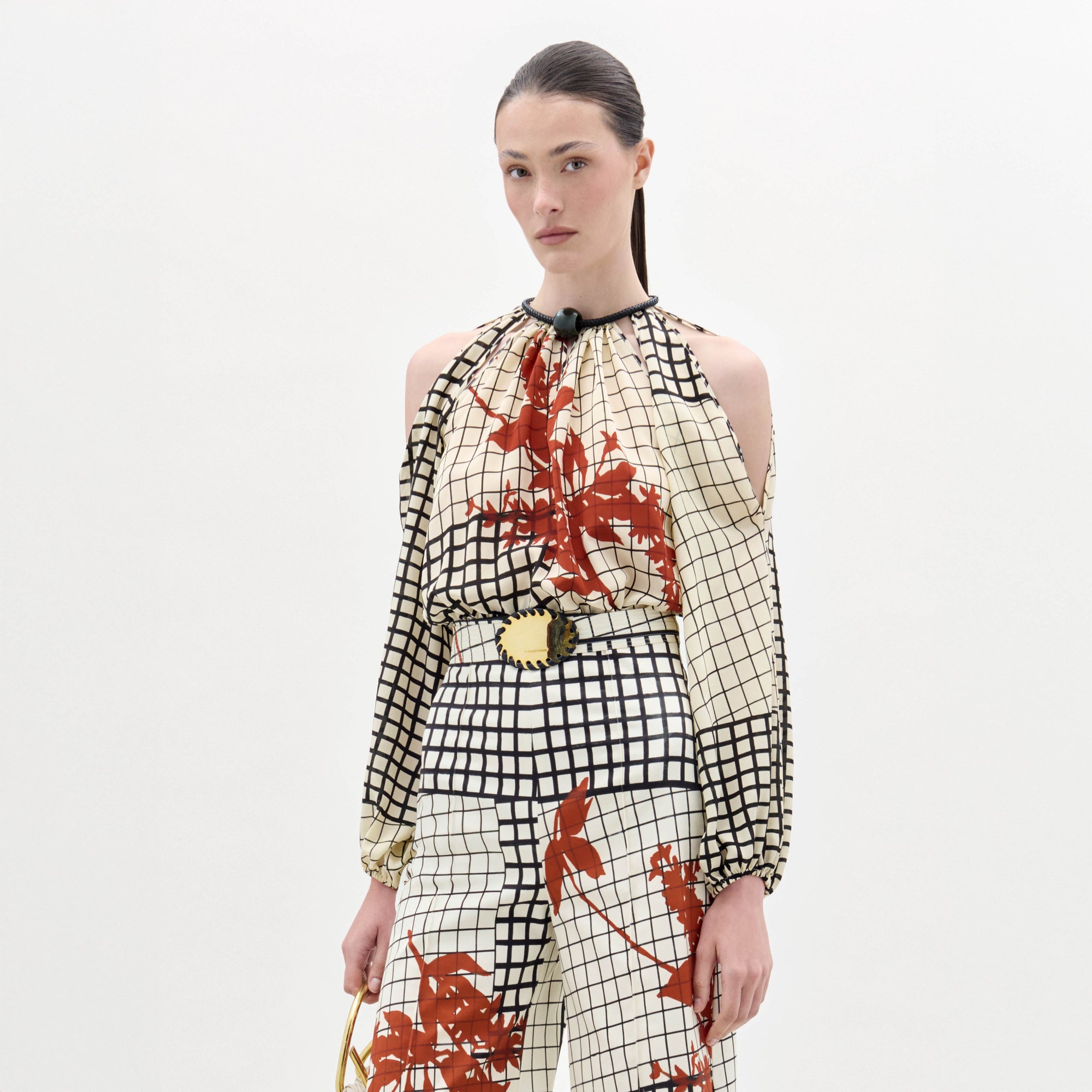 A woman wears the Elke Blouse, a cream and black grid-patterned top with red floral accents, cold-shoulder sleeves, and a belt with a circular gold buckle, standing against a plain white background.