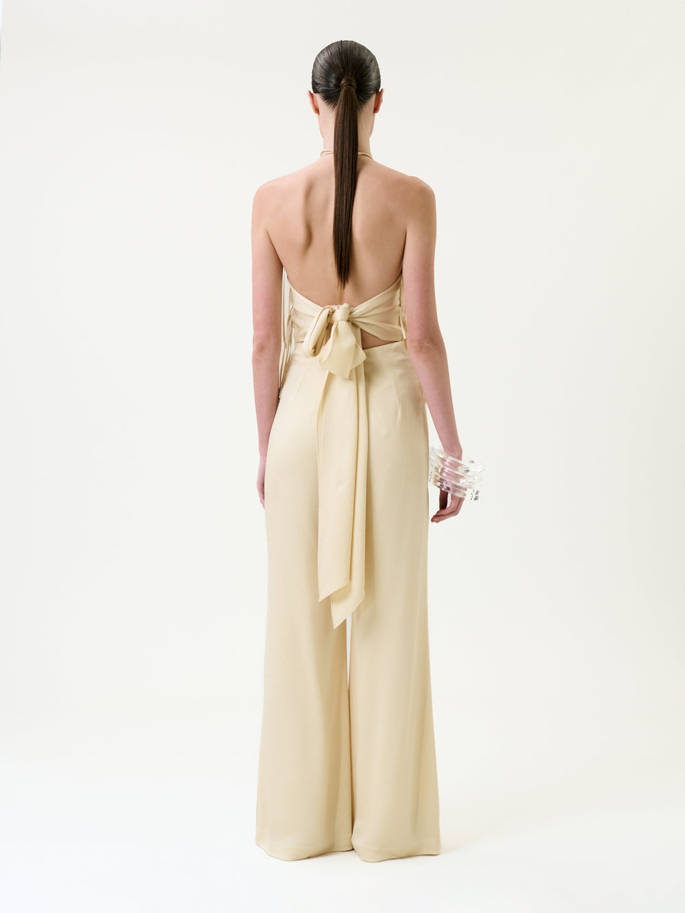 Woman with long dark hair in a low ponytail, wearing the backless Tulsi Jumpsuit in beige, featuring a large bow at the waist, stands facing away on a white background, holding clear bangles in one hand.