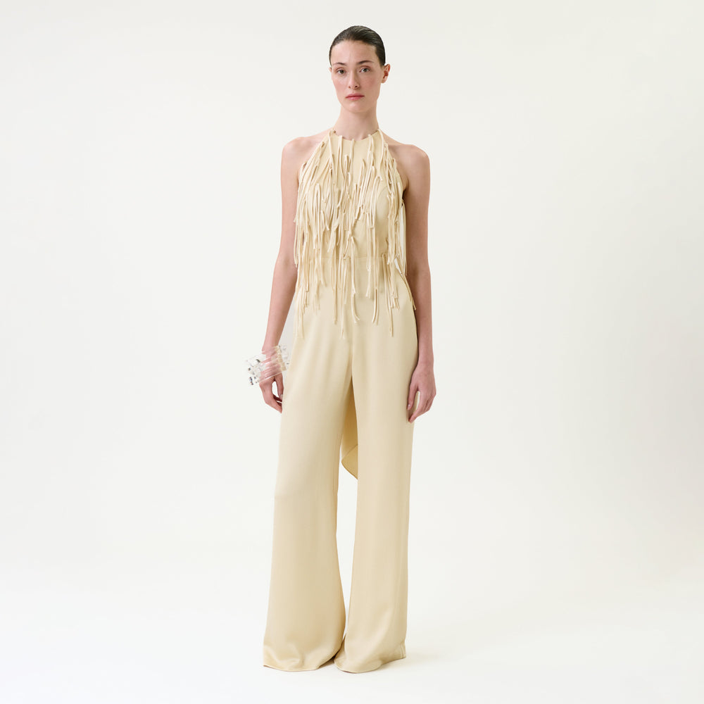 A woman stands against a plain white background wearing the Tulsi Jumpsuit, a cream-colored halter style with long fringe details on the bodice and wide-leg pants, paired with a white floral bracelet.