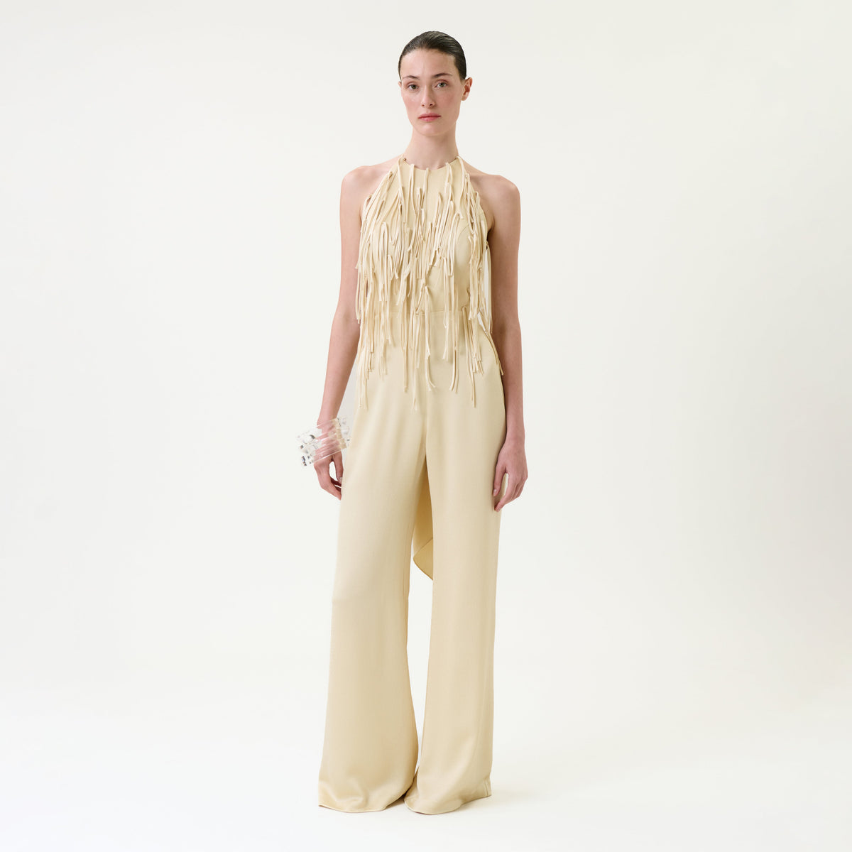 A woman stands against a plain white background wearing the Tulsi Jumpsuit, a cream-colored halter style with long fringe details on the bodice and wide-leg pants, paired with a white floral bracelet.