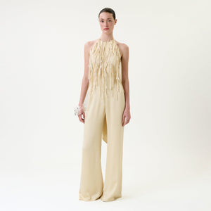 A woman stands against a plain white background wearing the Tulsi Jumpsuit, a cream-colored halter style with long fringe details on the bodice and wide-leg pants, paired with a white floral bracelet.