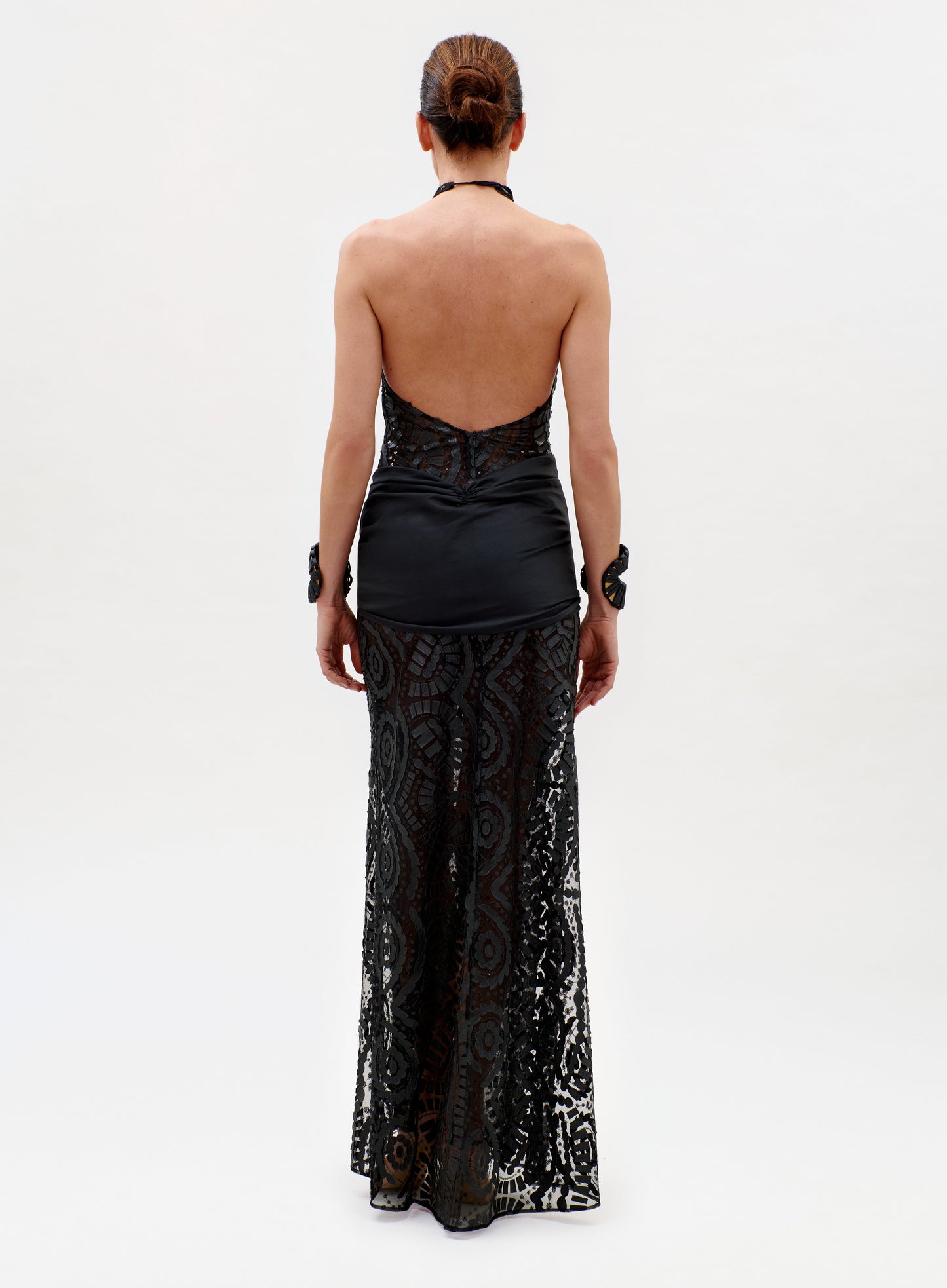 A person, facing away, wears the Lili Dress—a black halter-neck style with a sheer lace skirt, satin fitted top, open back, and two matching lace cuffs—against a plain white background.