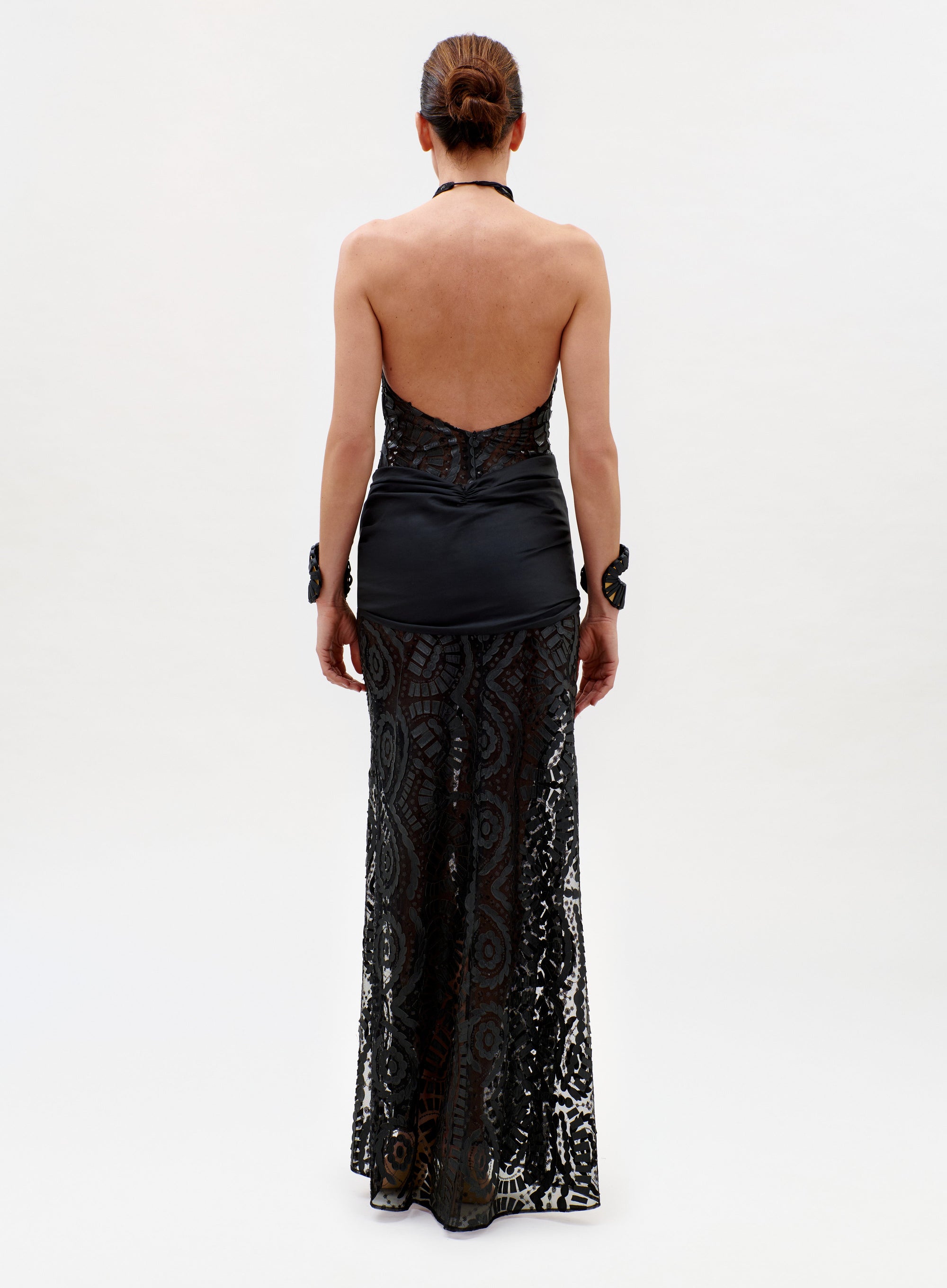 A person, facing away, wears the Lili Dress—a black halter-neck style with a sheer lace skirt, satin fitted top, open back, and two matching lace cuffs—against a plain white background.