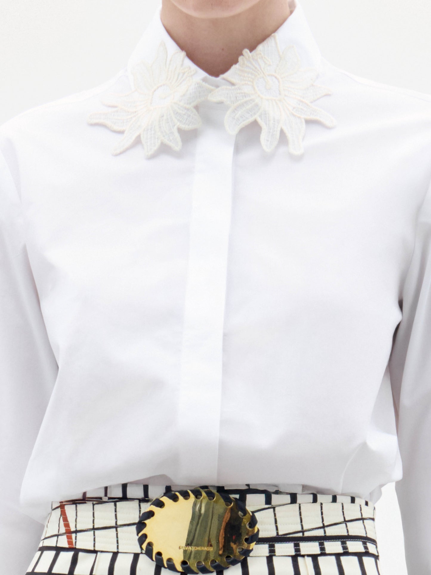 A woman stands against a white background wearing the Naroa Blouse, paired with high-waisted grid-and-floral print pants and a wide belt featuring a gold buckle.