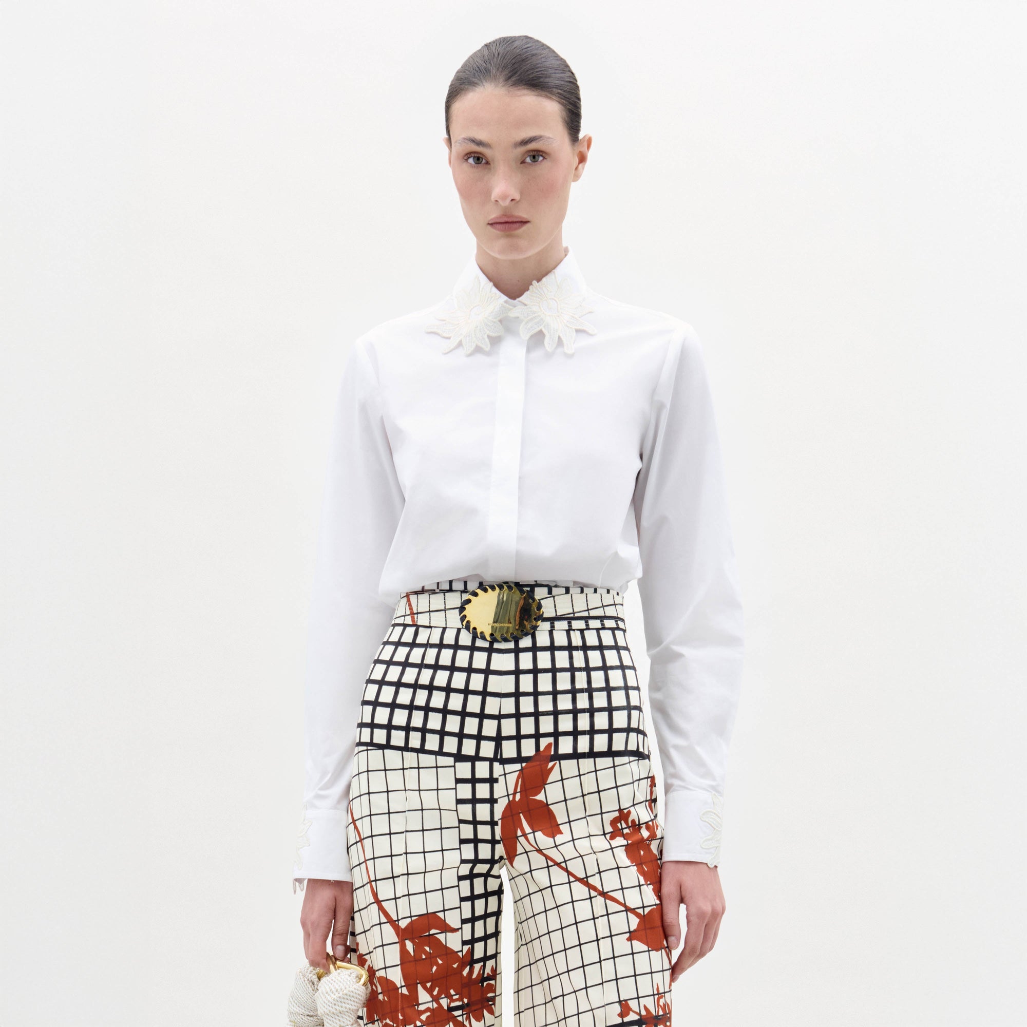 A woman stands against a white background wearing the Naroa Blouse, paired with high-waisted grid-and-floral print pants and a wide belt featuring a gold buckle.