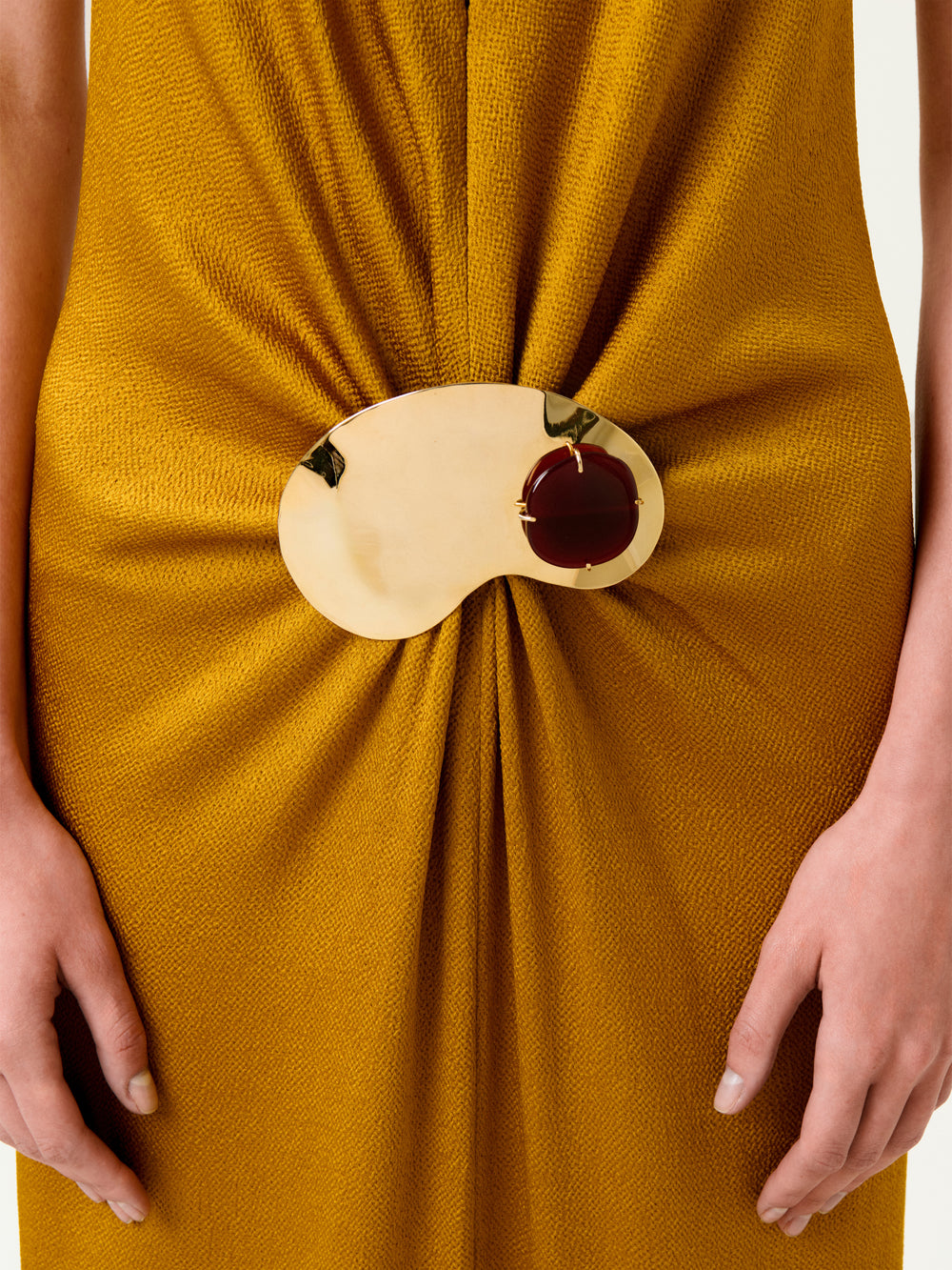 A person wearing the Sira Dress in mustard yellow, featuring a textured Italian fabric with the material elegantly gathered at the waist by a gold and brown bean-shaped decorative brooch. Only the waist and hands are visible.