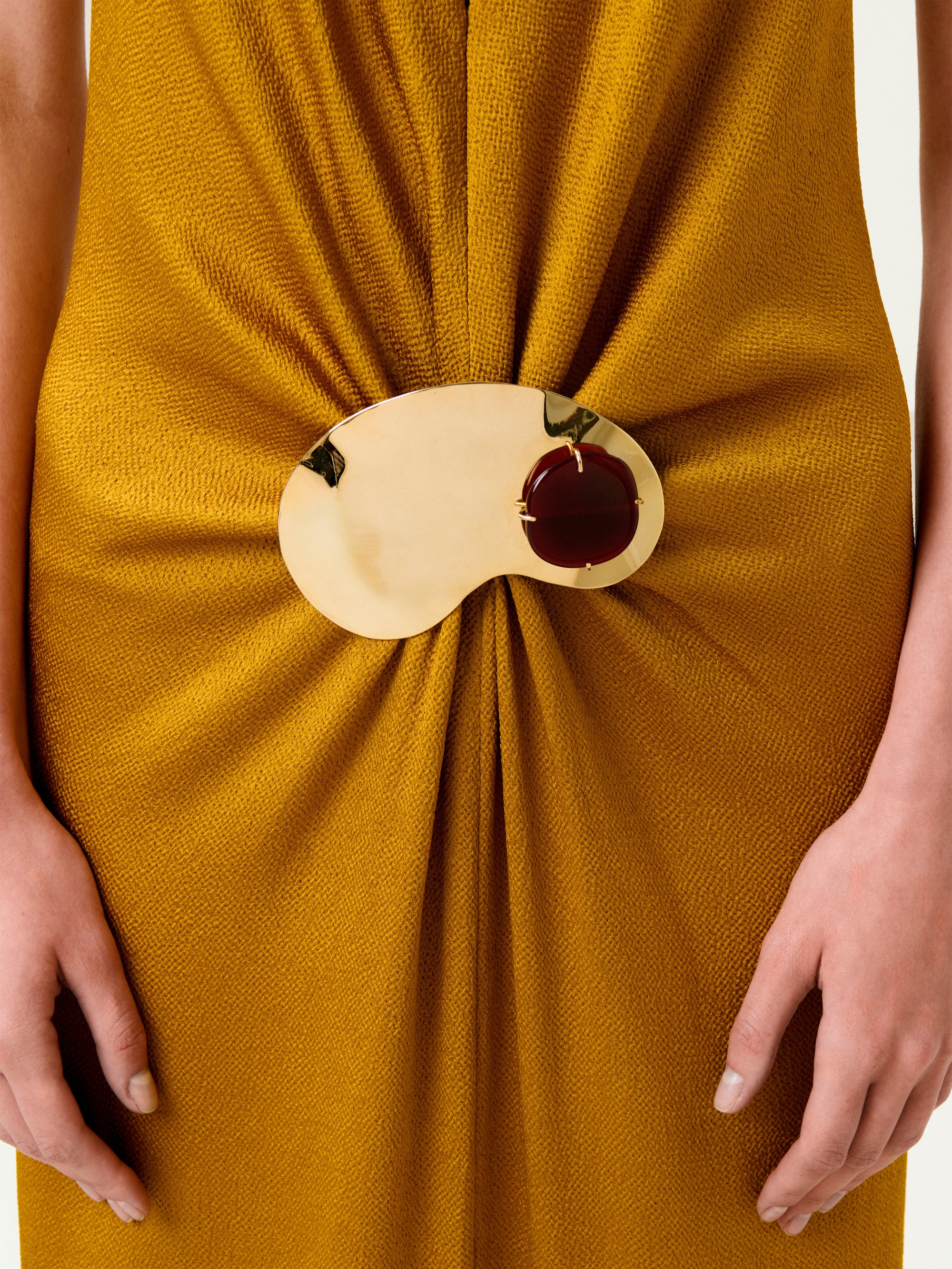 A person wearing the Sira Dress in mustard yellow, featuring a textured Italian fabric with the material elegantly gathered at the waist by a gold and brown bean-shaped decorative brooch. Only the waist and hands are visible.