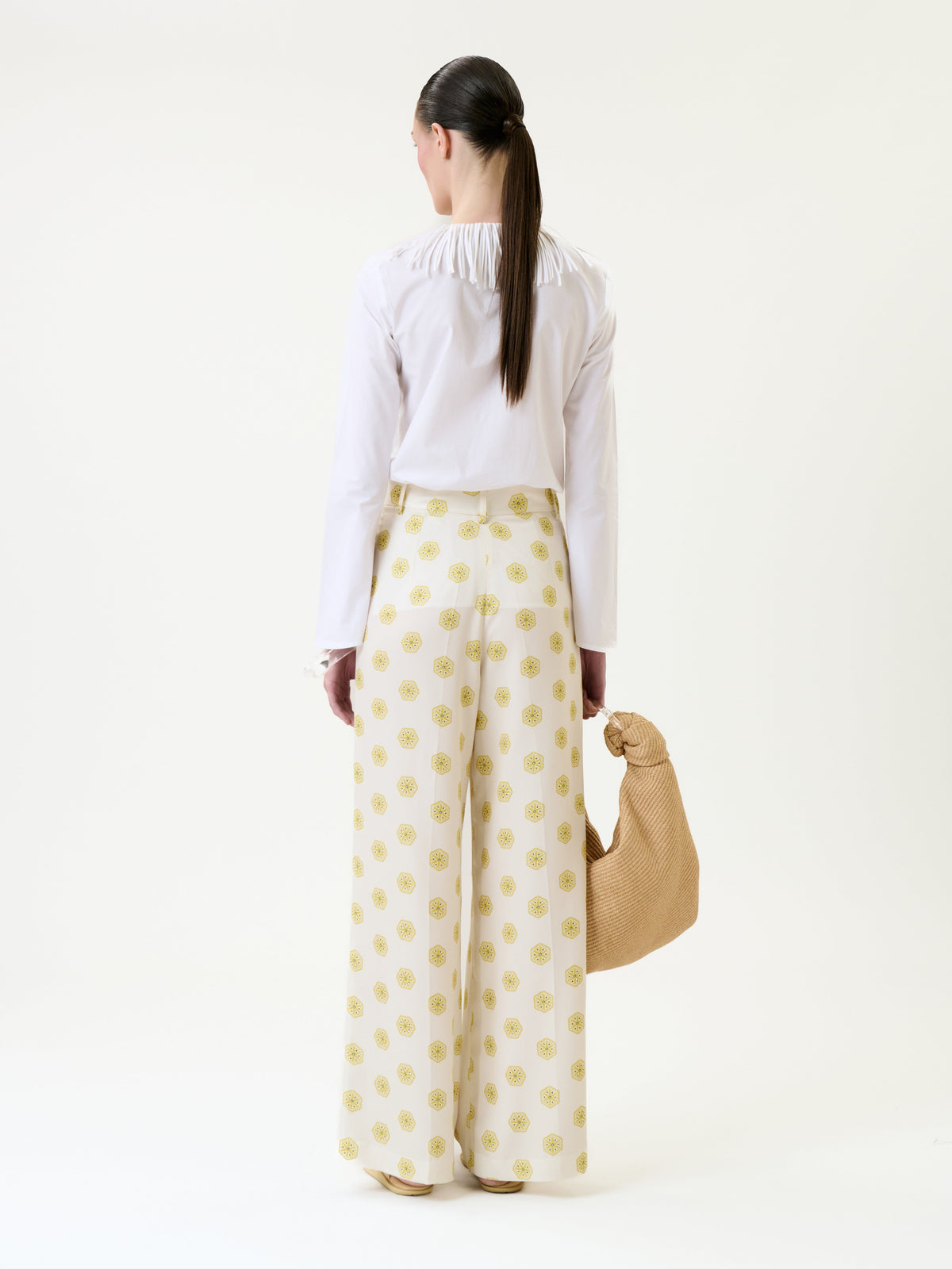 A person stands facing away, wearing a white blouse paired with the Emine Pant—wide-leg white pants featuring a yellow circular pattern—and holding a tan woven bag.