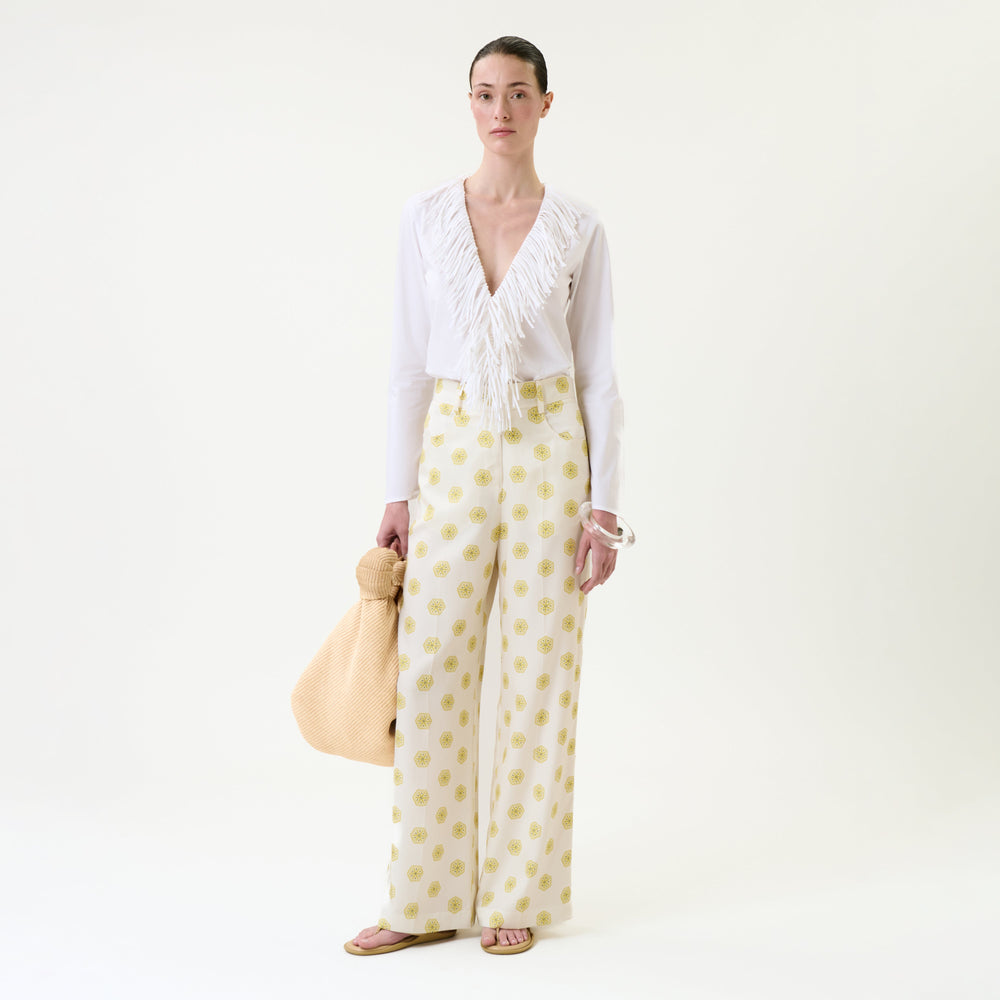 A woman stands against a plain background wearing a white blouse with a deep V and fringe detail, the Emine Pant – high-waisted wide-leg trousers featuring a yellow dot pattern, pink sandals, and holding a large beige textured bag.