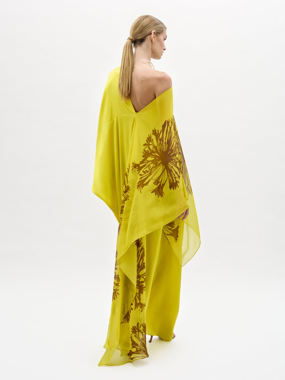 A woman stands against a plain white background wearing the Jade Dress, a yellow one-shoulder silk maxi with a bold brown floral print, paired with open-toe sandals.