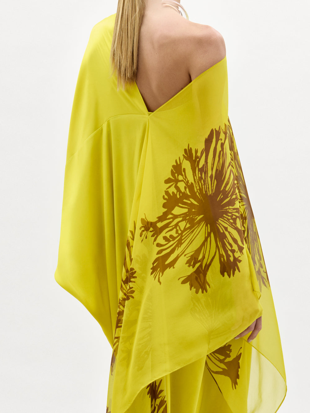 A woman stands against a plain white background wearing the Jade Dress, a yellow one-shoulder silk maxi with a bold brown floral print, paired with open-toe sandals.
