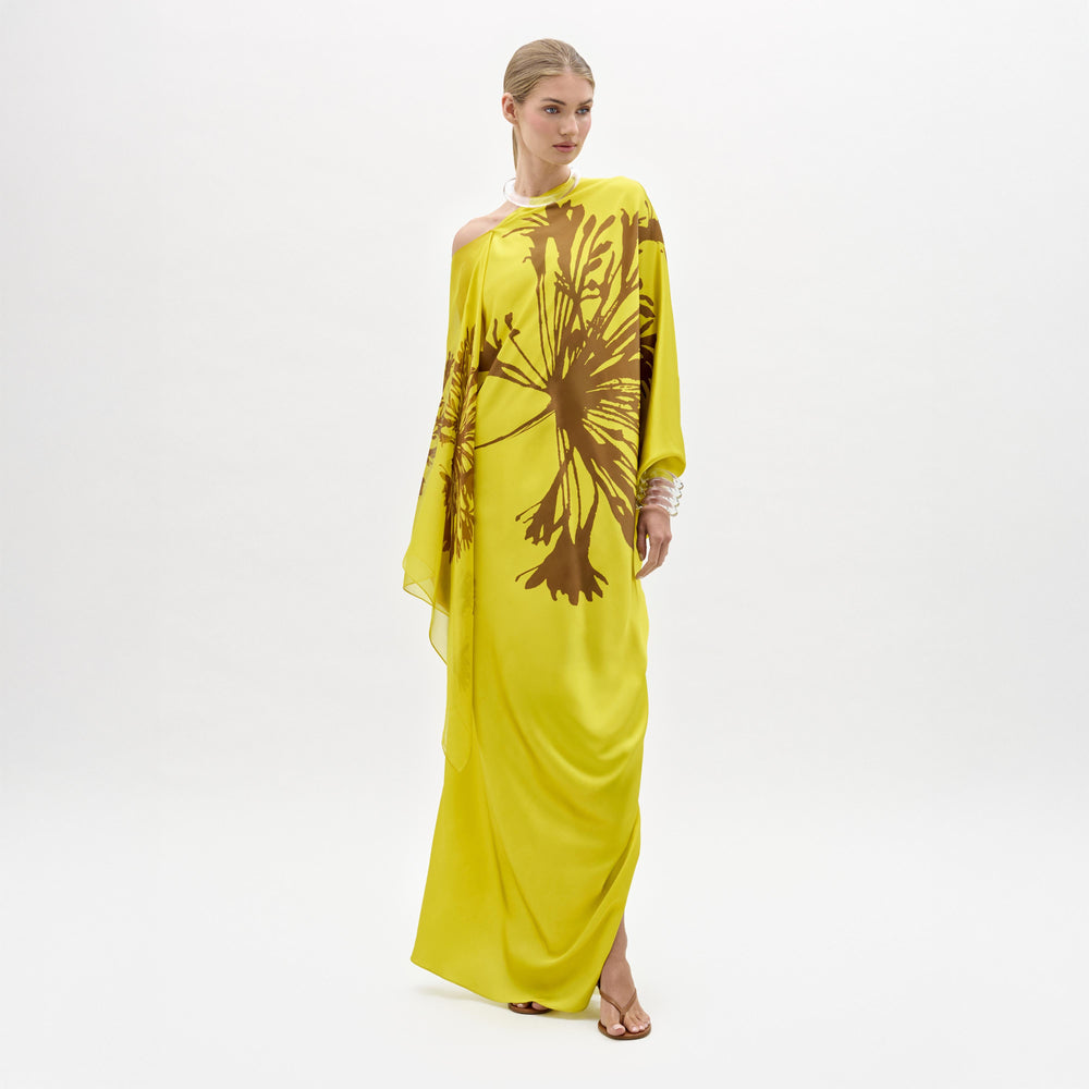 A woman stands against a plain white background wearing the Jade Dress, a yellow one-shoulder silk maxi with a bold brown floral print, paired with open-toe sandals.