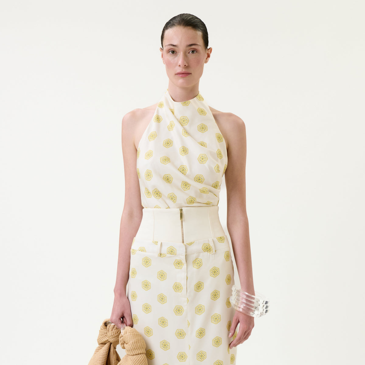 A woman with slicked-back hair wears the sleeveless, white Marine Blouse halter top and a matching skirt adorned with yellow circular patterns. She holds a textured beige garment and accessorizes with clear bangles on her wrist. The background is plain white.