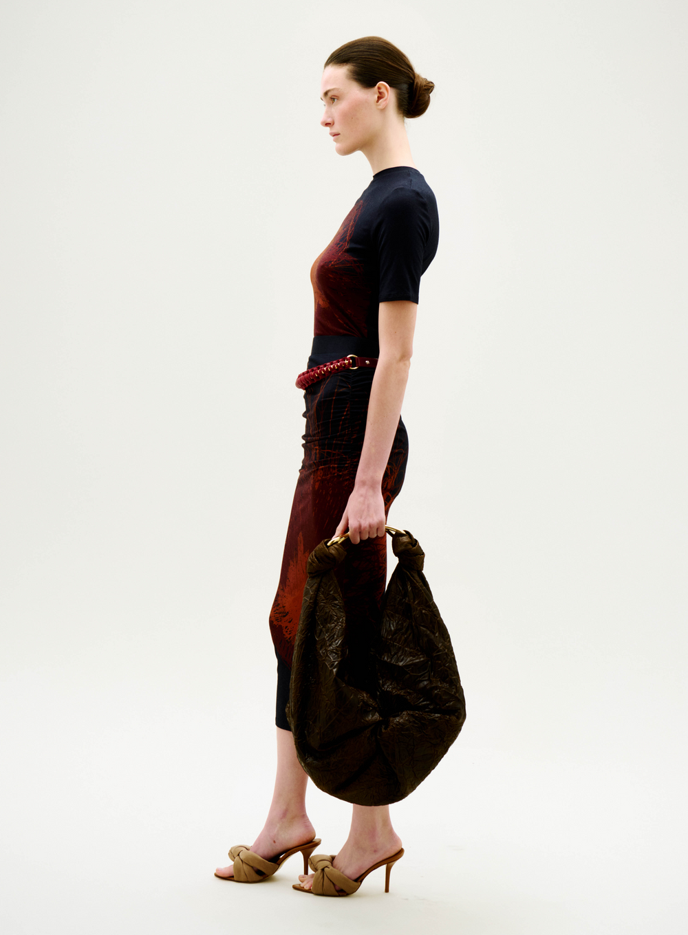 A woman in a fitted, patterned Niove Skirt and heeled sandals stands in profile, holding a large, textured brown bag against a plain white background.