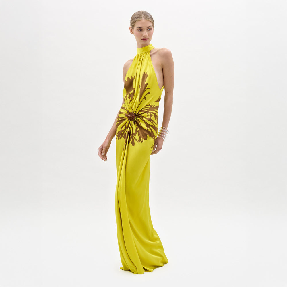A woman poses against a white background in the Koori Dress—a sleeveless, floor-length yellow halter maxi with a brown floral pattern, minimal jewelry, and a draped column silhouette.