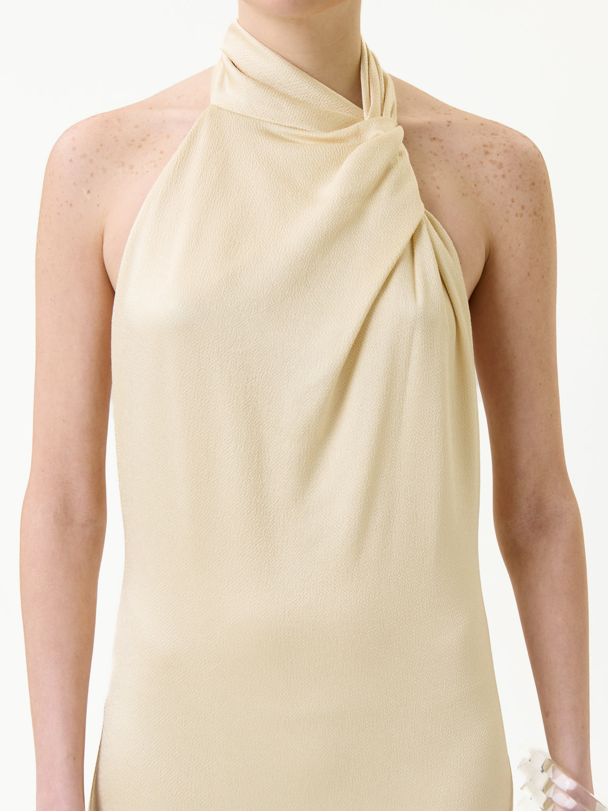 A person wearing the Maica Dress—a sleeveless, beige, halter-neck style with draped fabric—standing against a plain light background.