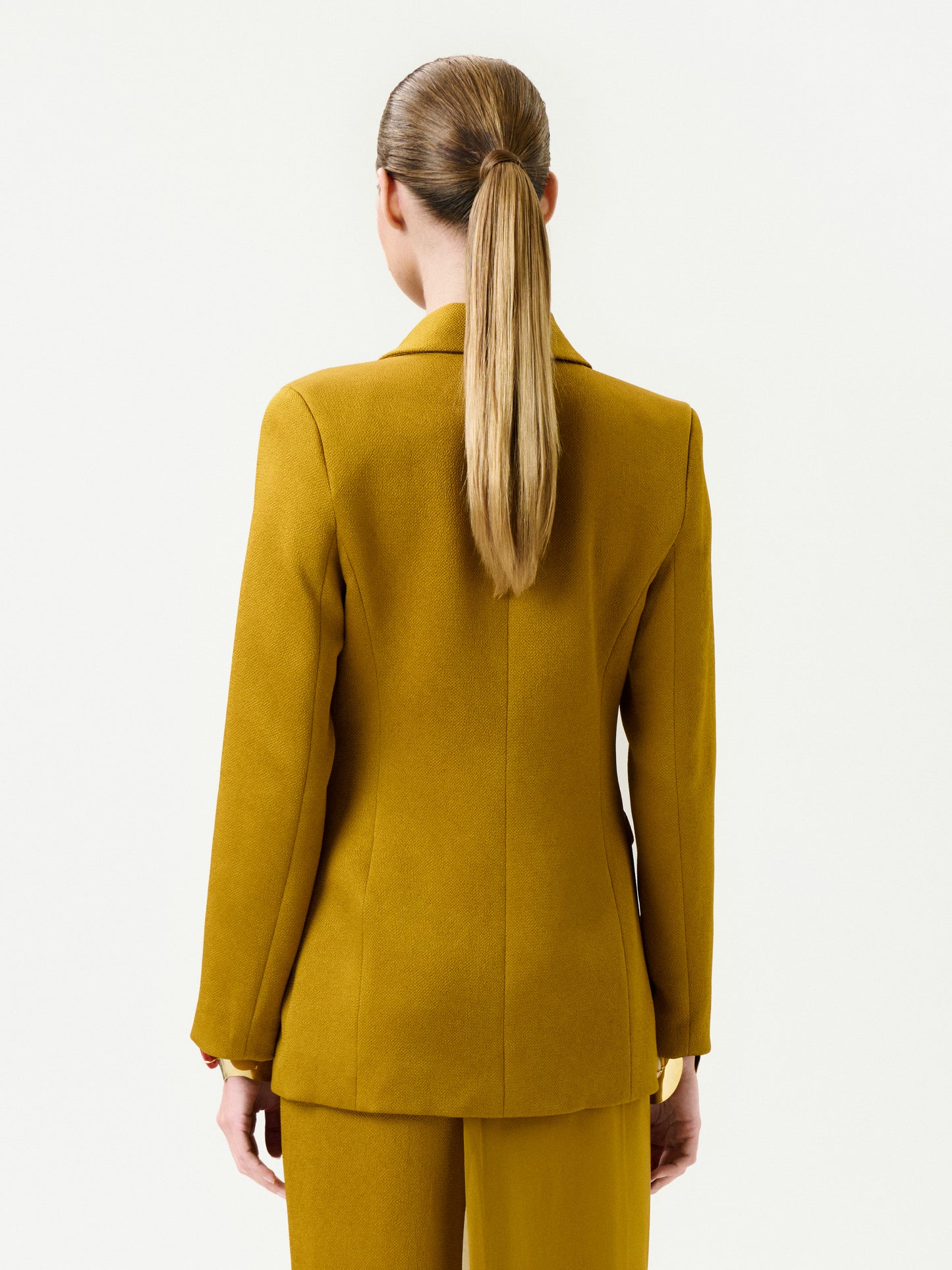 A woman with straight, light brown hair in a low ponytail is seen from the back, wearing the mustard yellow Rebeca Jacket and matching pants against a plain white background.