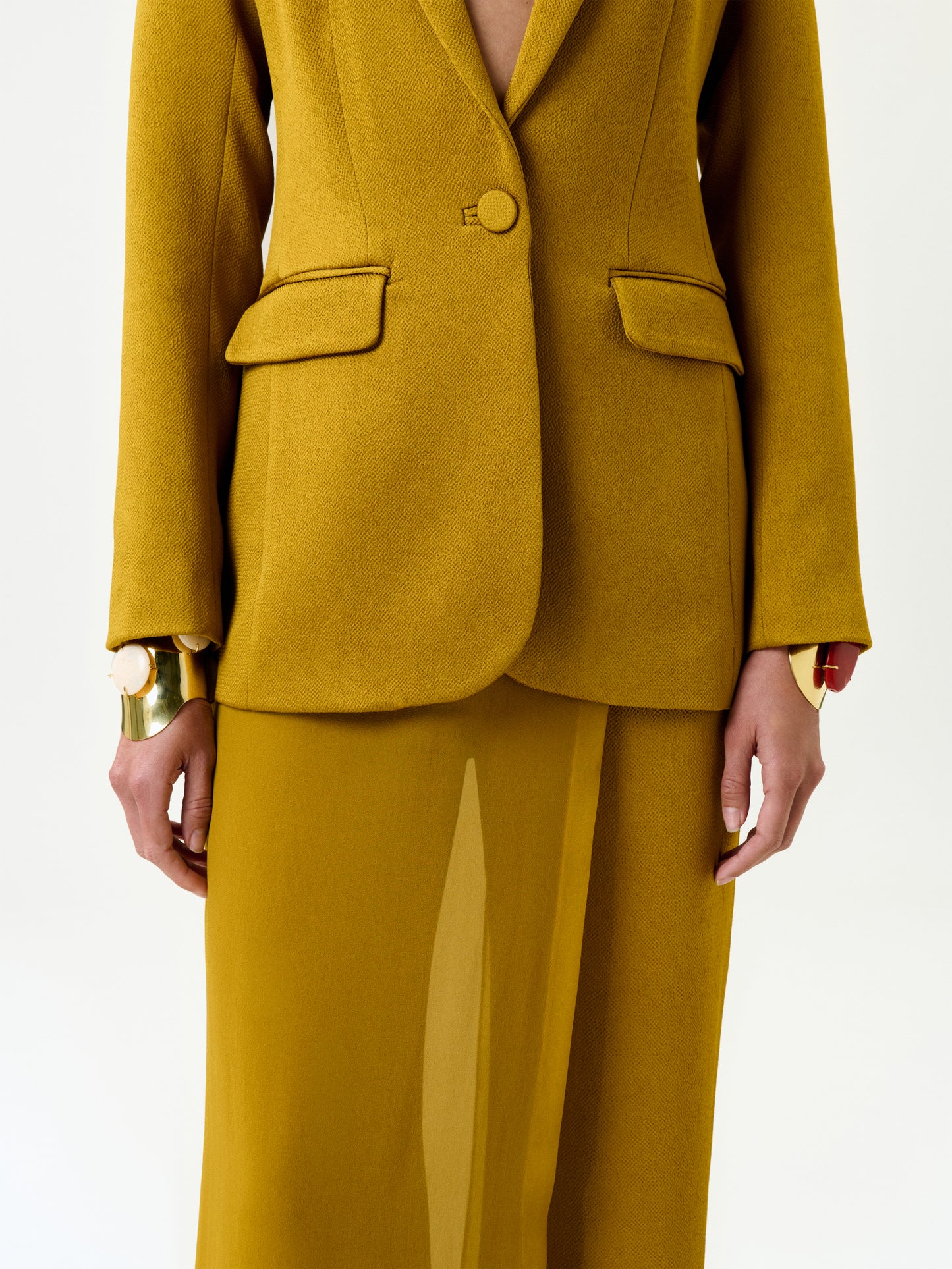 A model wears the Rebeca Jacket in mustard yellow Italian fabric with a matching skirt and gold statement bracelets, shown from shoulders to knees against a plain white background.