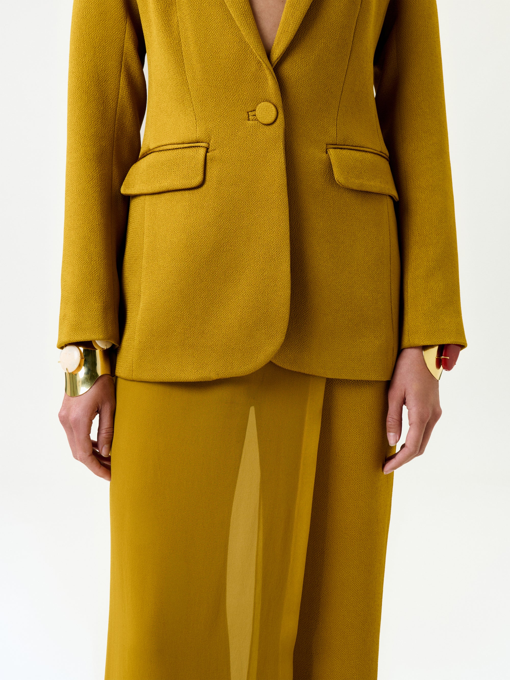 A model wears the Rebeca Jacket in mustard yellow Italian fabric with a matching skirt and gold statement bracelets, shown from shoulders to knees against a plain white background.