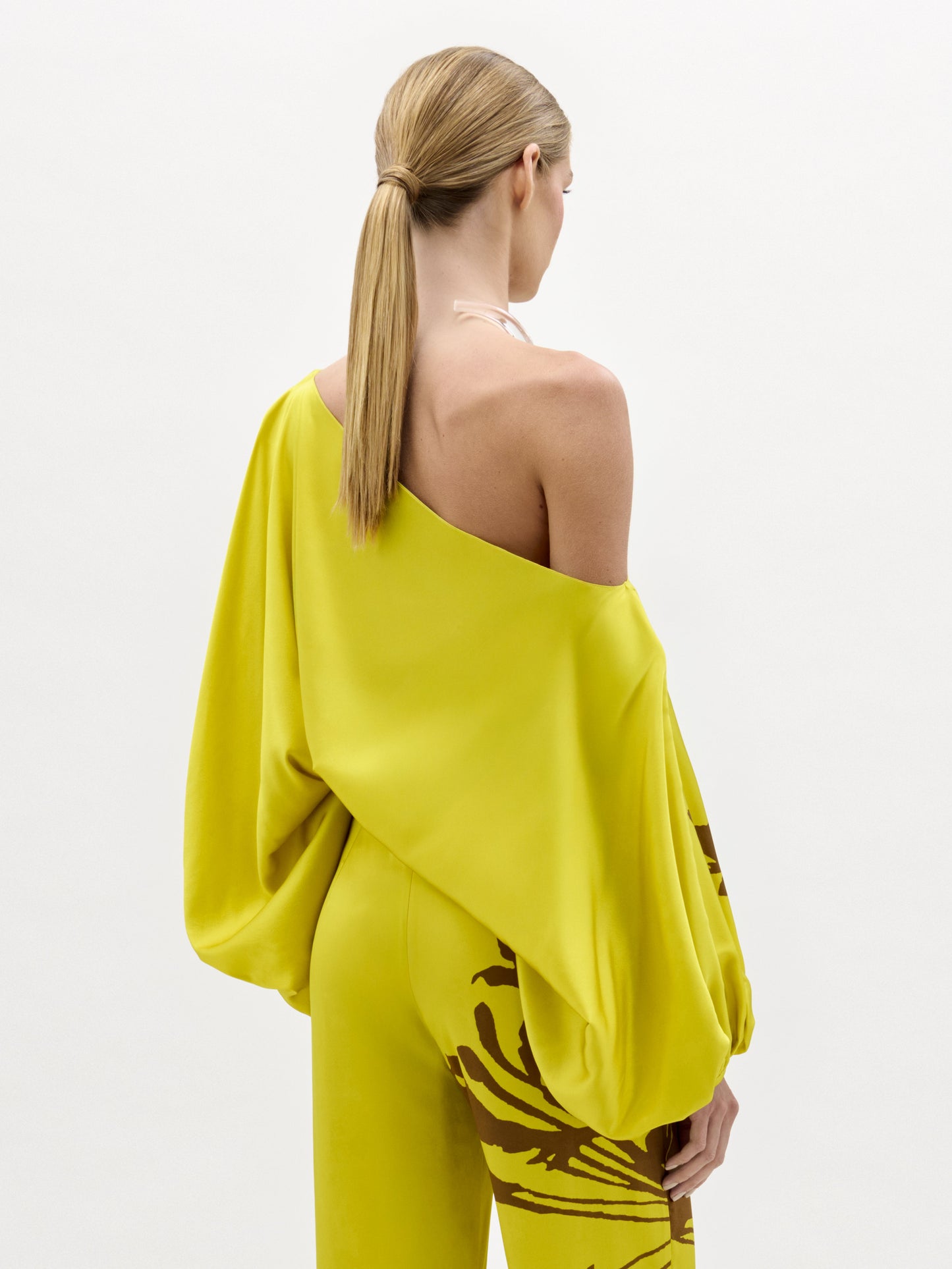 A woman models the Bellagio Blouse—a yellow silk, one-shoulder top featuring brown abstract patterns and draped sleeves—standing against a plain white background.
