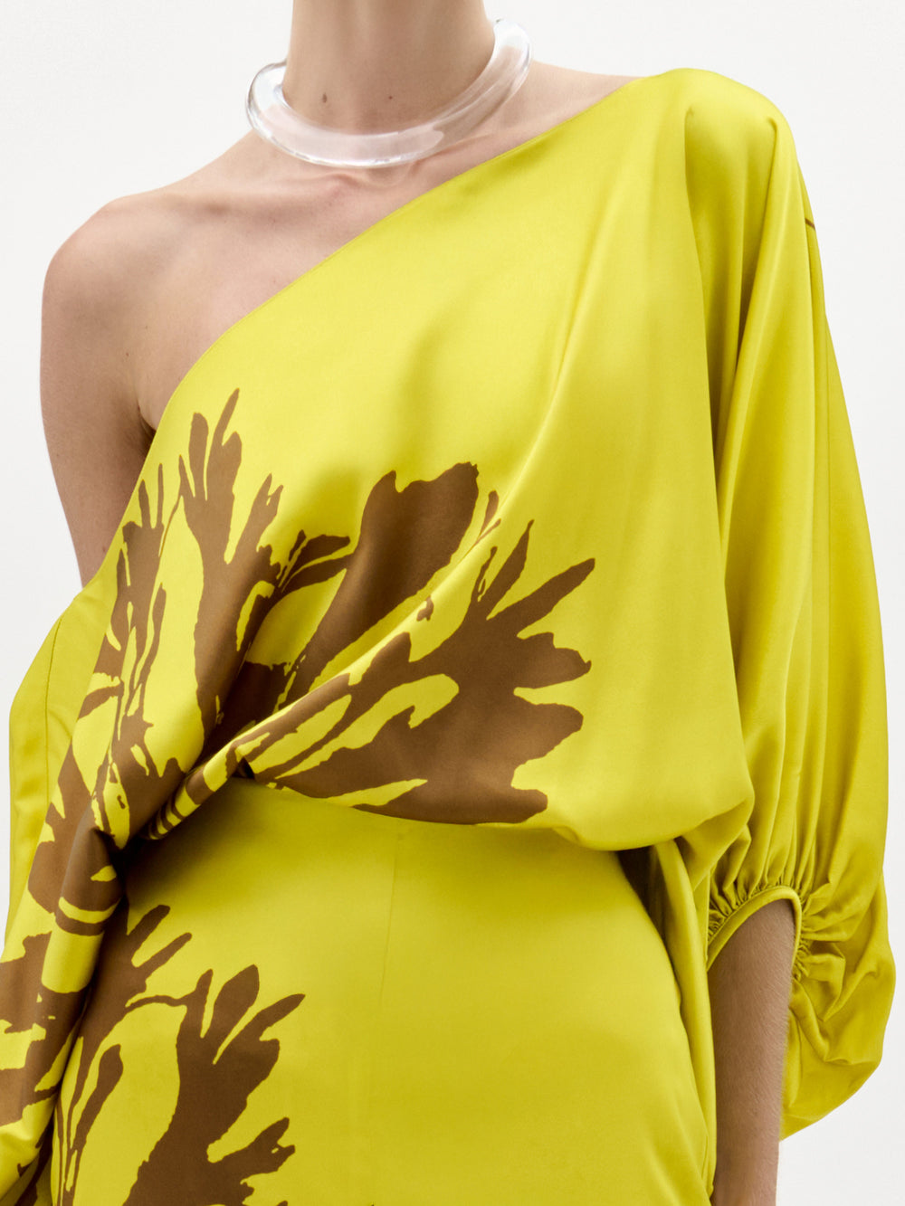 A woman models the Bellagio Blouse—a yellow silk, one-shoulder top featuring brown abstract patterns and draped sleeves—standing against a plain white background.