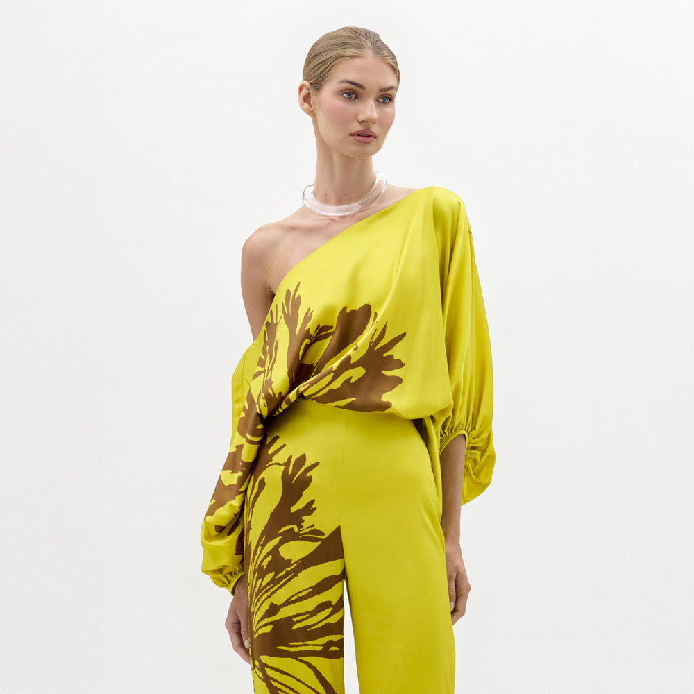 A woman models the Bellagio Blouse—a yellow silk, one-shoulder top featuring brown abstract patterns and draped sleeves—standing against a plain white background.