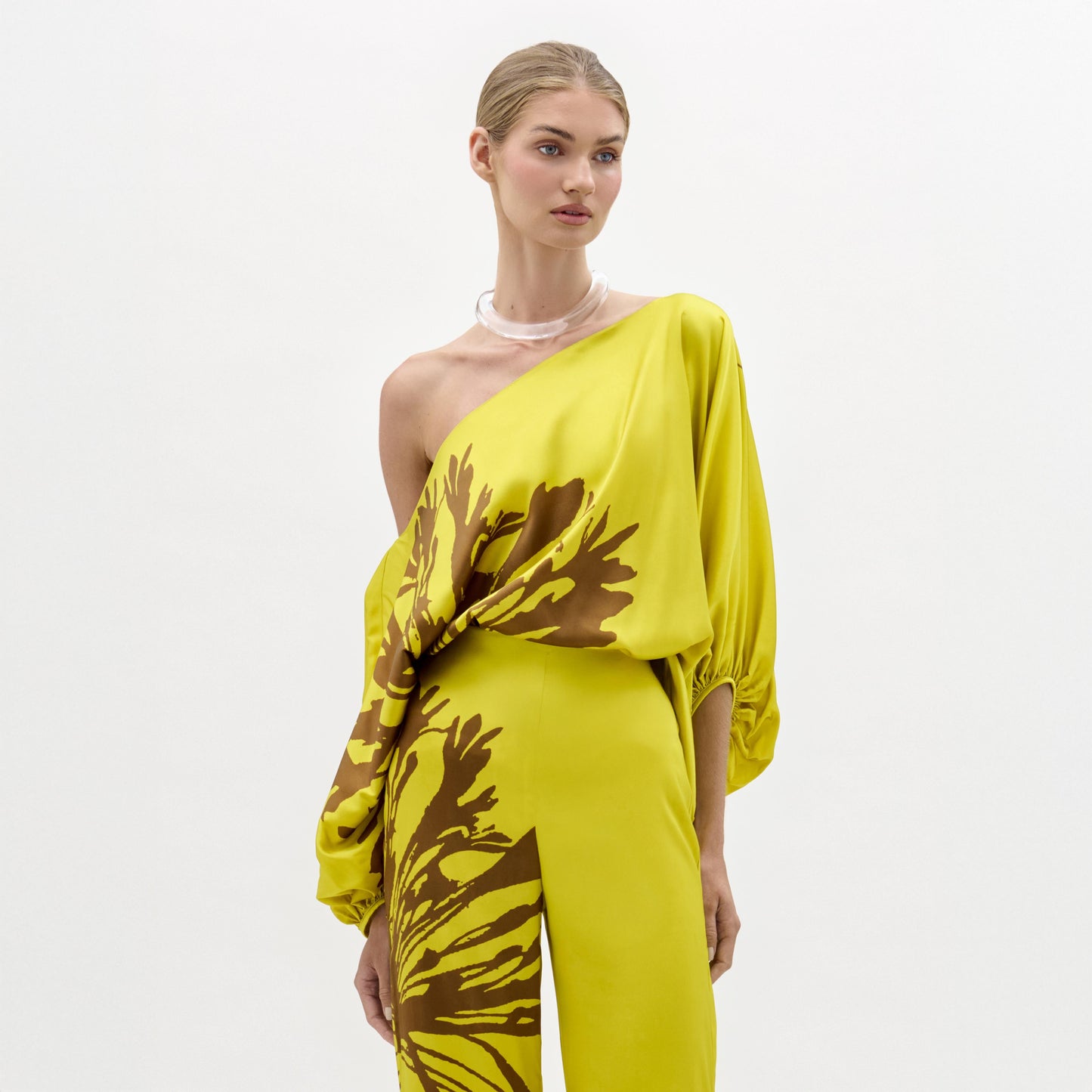 A woman models the Bellagio Blouse—a yellow silk, one-shoulder top featuring brown abstract patterns and draped sleeves—standing against a plain white background.