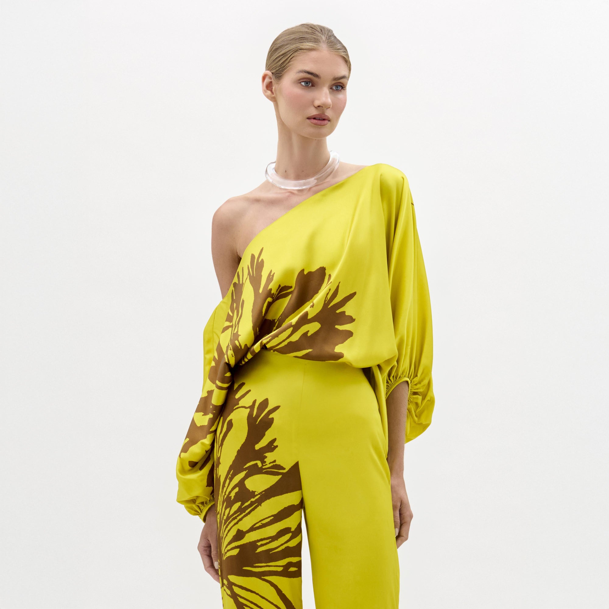 A woman models the Bellagio Blouse—a yellow silk, one-shoulder top featuring brown abstract patterns and draped sleeves—standing against a plain white background.