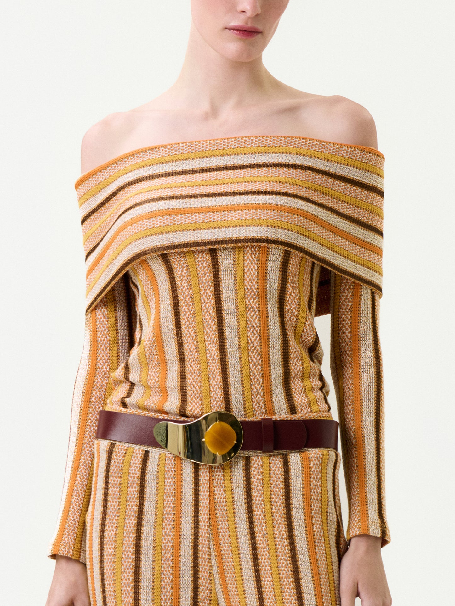 A woman wears the Maitenea Top—an off-the-shoulder, long-sleeved knit with vertical yellow and brown stripes—paired with matching pants and a wide brown belt with a gold buckle. Made in Colombia with a true-to-size fit.