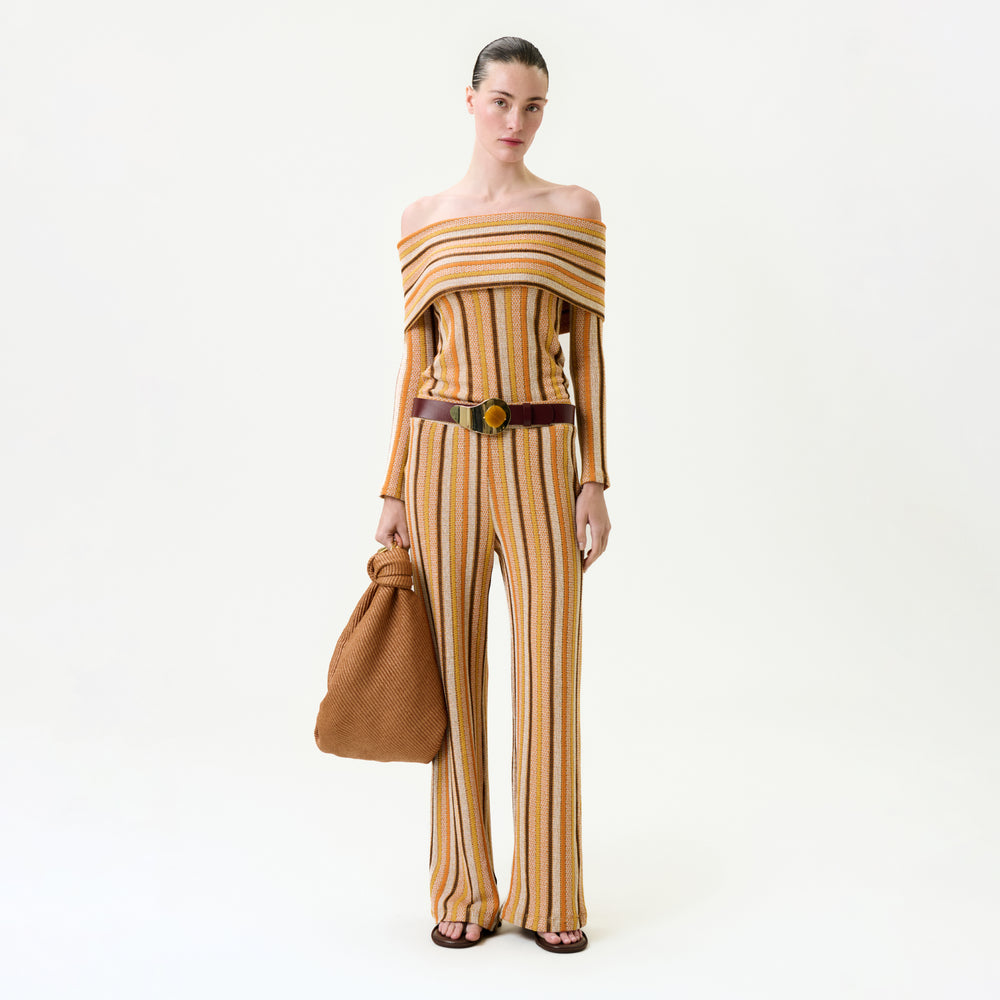A woman stands against a white background wearing the Naoki Pant, an off-the-shoulder striped knit jumpsuit in yellow, orange, and beige. She accessorizes with a large brown textured bag, wide brown belt, and sandals. Fits true to size.