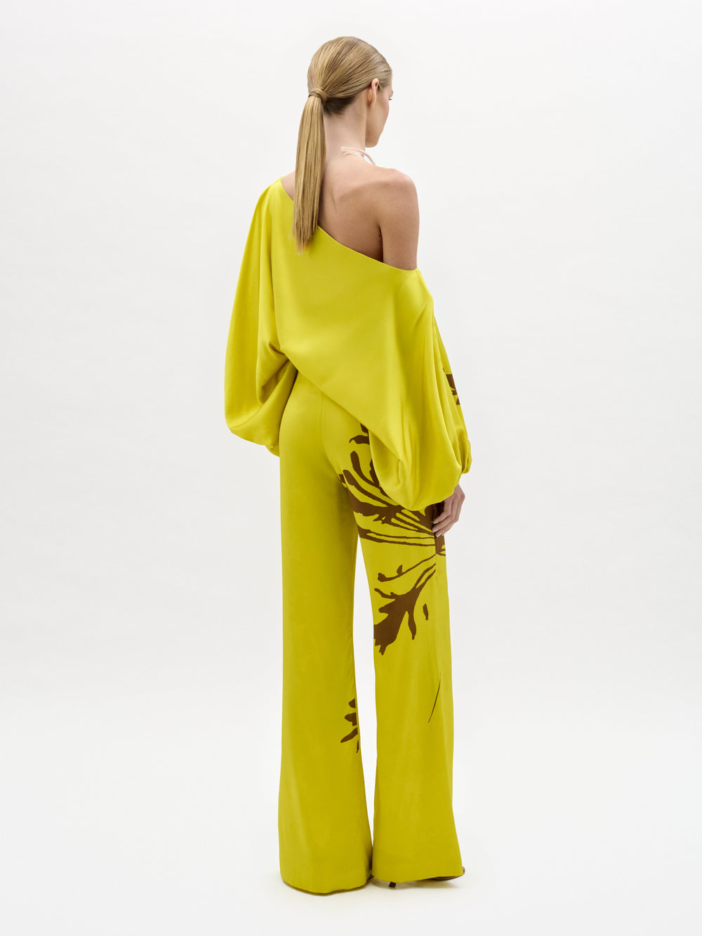 A woman stands against a plain background wearing the Andie Pant, a wide-leg, one-shoulder yellow jumpsuit with brown floral designs and a side slit. Made from luxe Italian fabric, she completes the look with sandals and a chunky necklace.