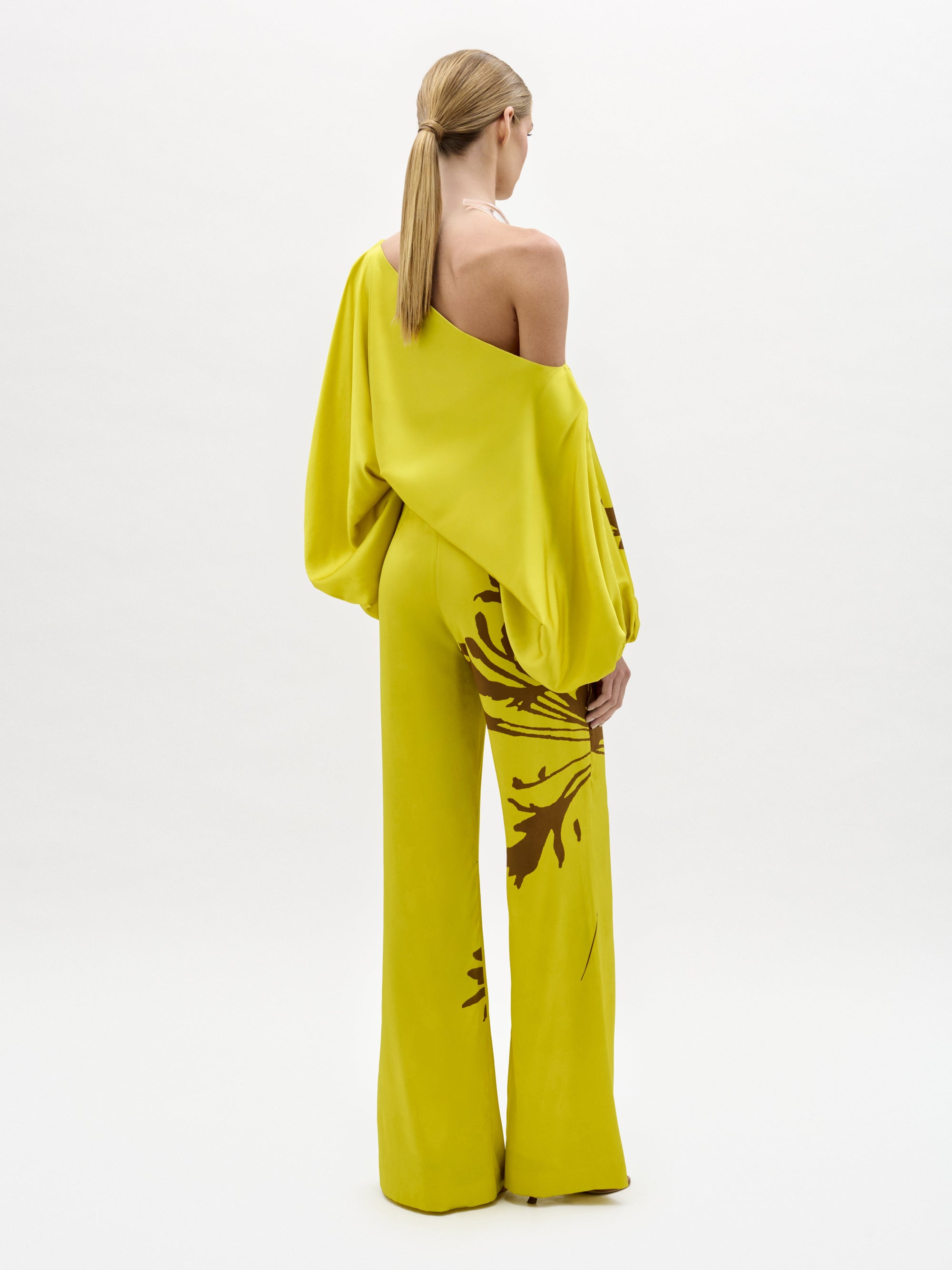 A woman stands against a plain background wearing the Andie Pant, a wide-leg, one-shoulder yellow jumpsuit with brown floral designs and a side slit. Made from luxe Italian fabric, she completes the look with sandals and a chunky necklace.
