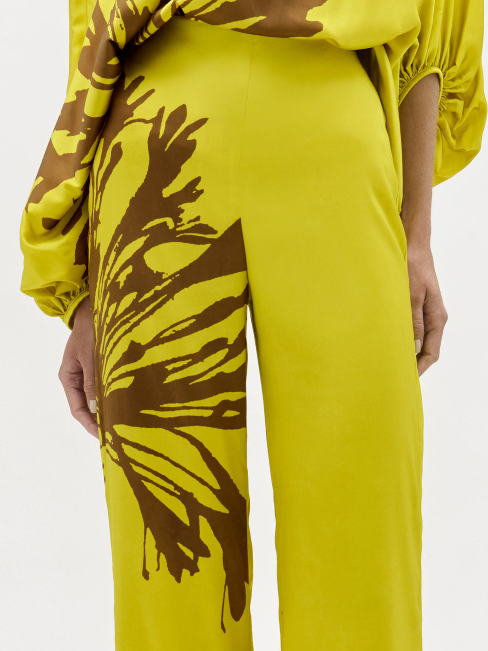 A woman stands against a plain background wearing the Andie Pant, a wide-leg, one-shoulder yellow jumpsuit with brown floral designs and a side slit. Made from luxe Italian fabric, she completes the look with sandals and a chunky necklace.