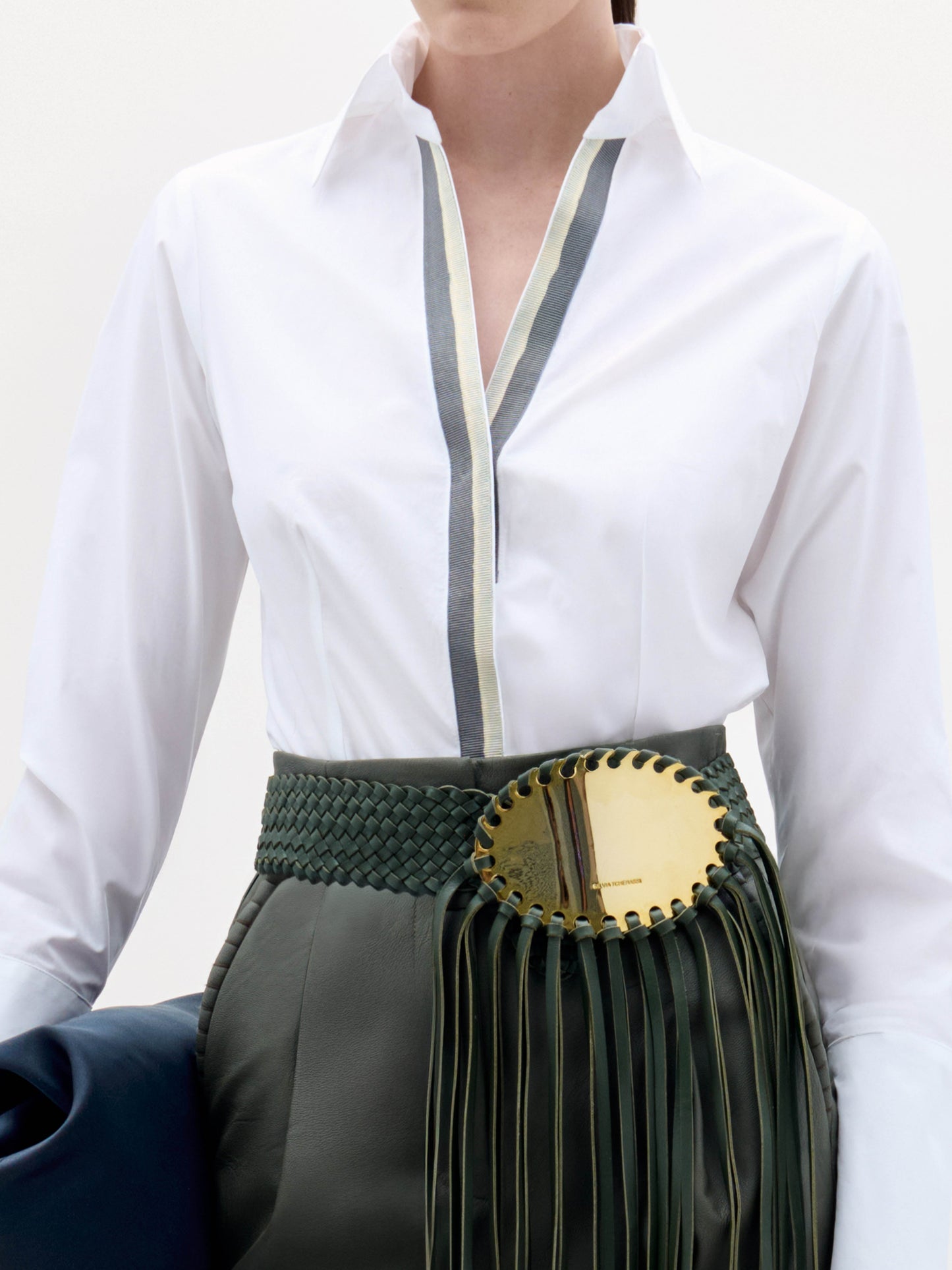 A woman stands against a white background wearing the Andrea Blouse, dark green belted pants with fringe detail, heeled sandals, and holding a dark clutch.