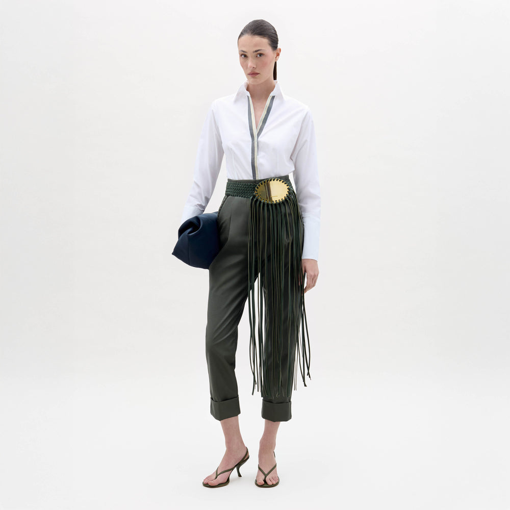 A woman stands against a white background wearing the Andrea Blouse, dark green belted pants with fringe detail, heeled sandals, and holding a dark clutch.