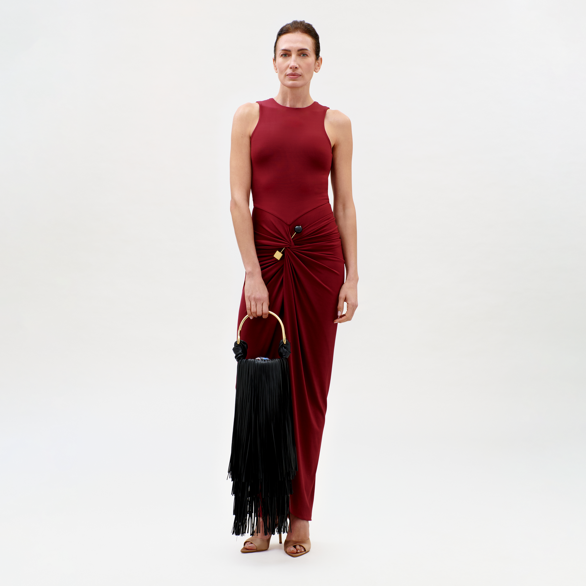 A woman wears the Leni Dress—sleeveless, floor-length, and burgundy with a knotted waist detail—styled with a black fringe handbag and tan open-toe heels. She stands against a simple light background.