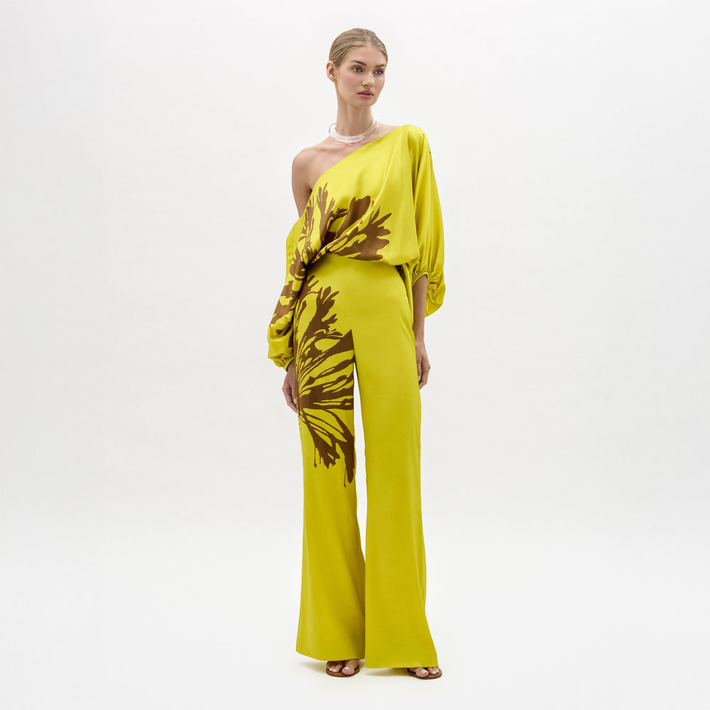 A woman stands against a plain background wearing the Andie Pant, a wide-leg, one-shoulder yellow jumpsuit with brown floral designs and a side slit. Made from luxe Italian fabric, she completes the look with sandals and a chunky necklace.