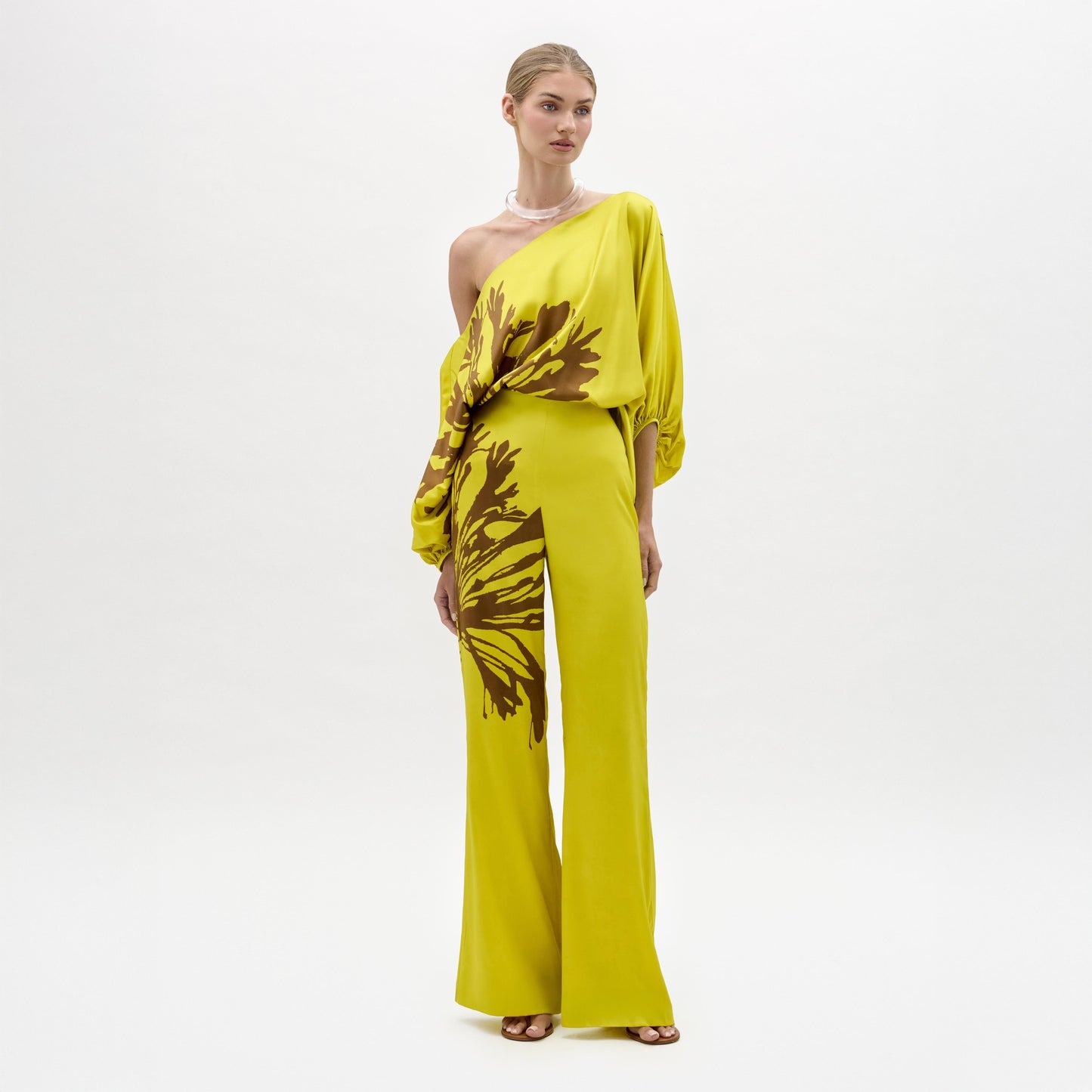A woman stands against a plain background wearing the Andie Pant, a wide-leg, one-shoulder yellow jumpsuit with brown floral designs and a side slit. Made from luxe Italian fabric, she completes the look with sandals and a chunky necklace.