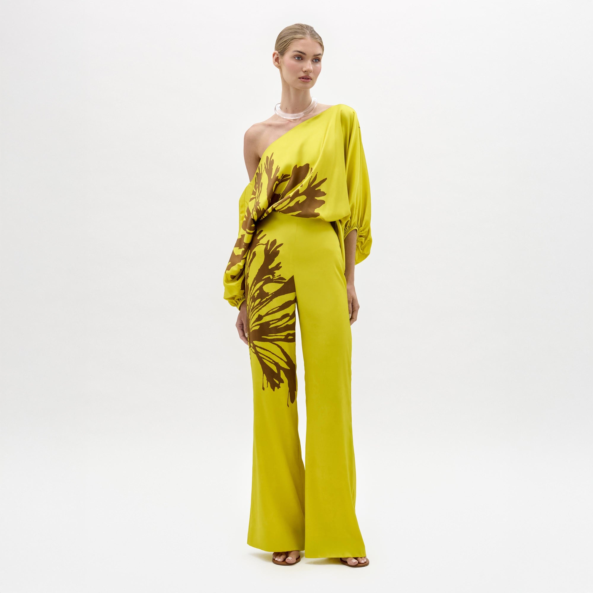 A woman stands against a plain background wearing the Andie Pant, a wide-leg, one-shoulder yellow jumpsuit with brown floral designs and a side slit. Made from luxe Italian fabric, she completes the look with sandals and a chunky necklace.