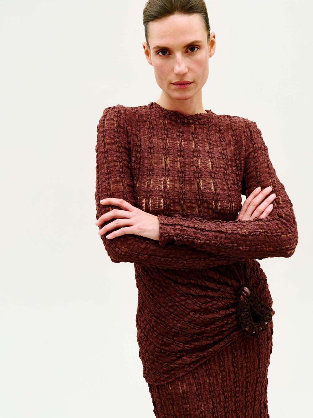 A woman with arms crossed wears the Roma T-Shirt, a textured, long-sleeved brown knit top, standing against a plain light background and looking directly at the camera with a neutral expression.