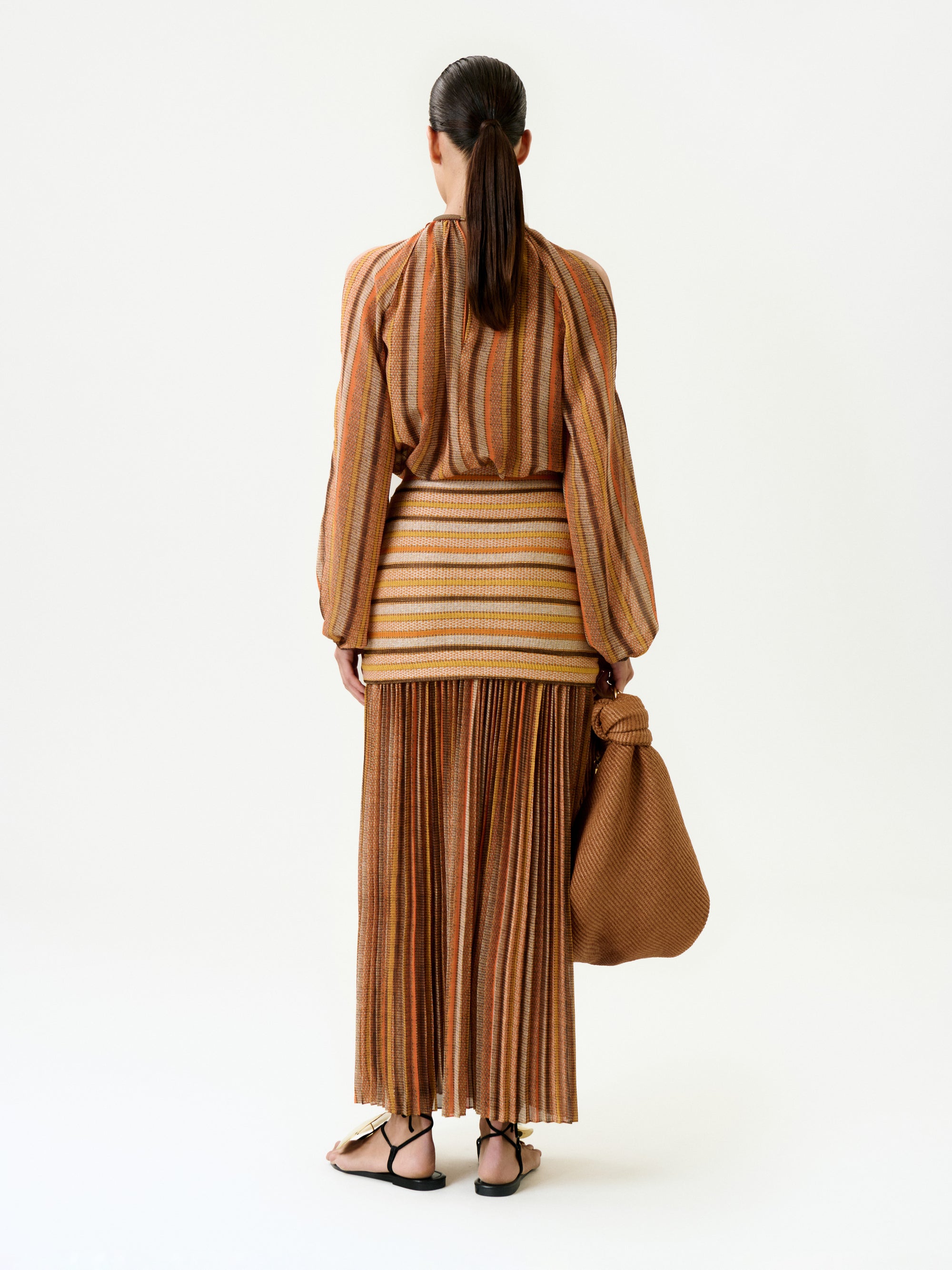 A woman with tied-back hair stands against a white background, wearing the Neko Skirt—a long, pleated, earth-toned striped skirt—paired with a wide-sleeved blouse, black sandals, and carrying a large woven brown bag.