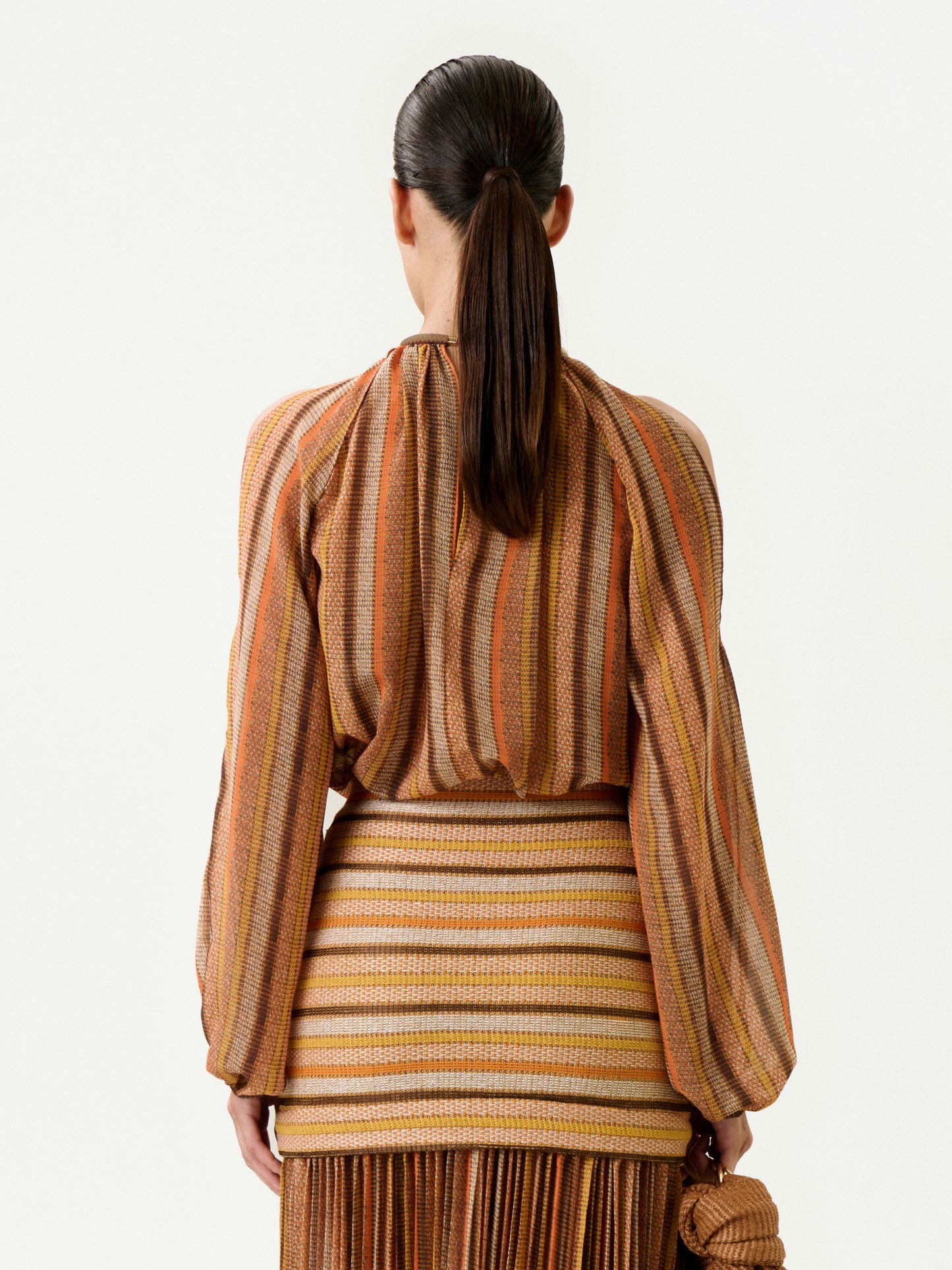 A woman with straight dark hair in a low ponytail is seen from behind, wearing the earth-toned striped Ona Blouse and a fitted skirt made from Italian fabric. She holds a small textured brown bag in her left hand.