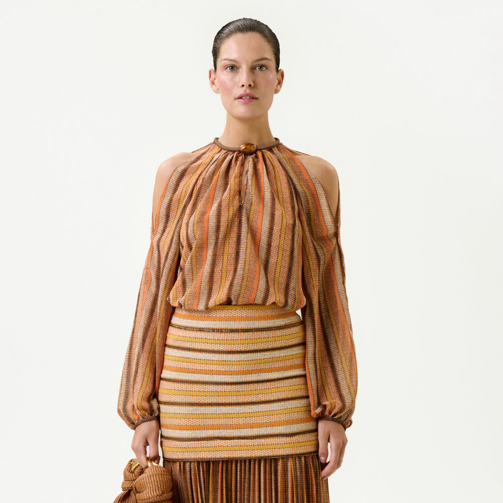 A woman stands facing forward, wearing the Ona Blouse—a long-sleeved, striped blouse with shoulder cutouts in orange, brown, and beige Italian fabric (100% Polyester)—paired with a matching skirt and holding a textured handbag against a white background.