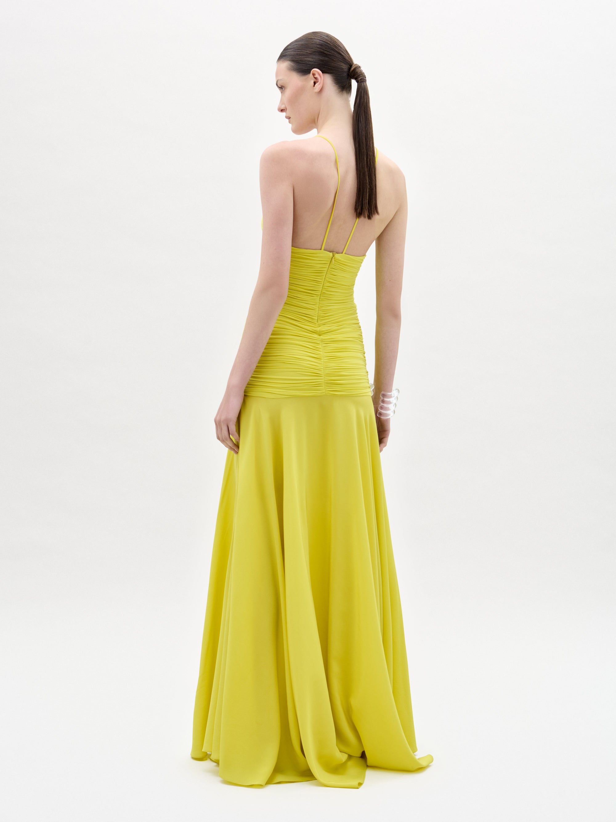 A woman poses against a plain white background wearing the Sana Dress, a sleeveless, floor-length yellow maxi with a ruched bodice, deep V neckline, and delicate floral appliqué.