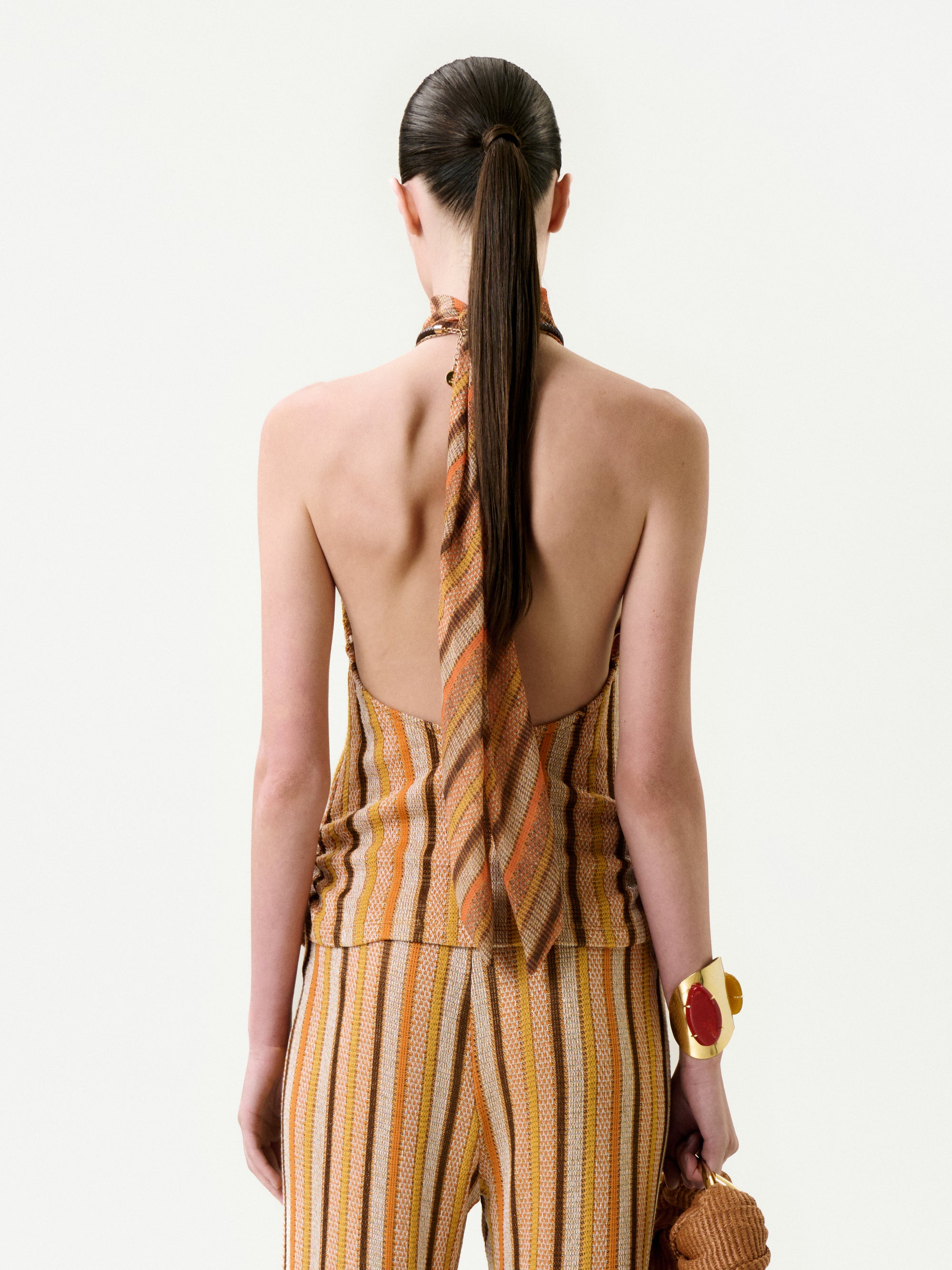 A person with long, dark hair in a ponytail is seen from behind wearing the Mitzy Blouse—a backless, striped halter top in warm tones made from Italian fabric—styled with matching pants, a chunky bracelet, and a textured bag.