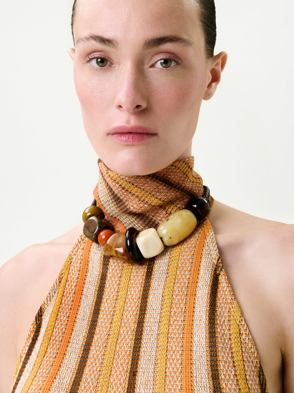 A woman with light skin and slicked-back hair wears the Mitzy Blouse—a sleeveless, high-neck top with orange, yellow, and brown stripes, a chunky beaded necklace, a true-to-size fit, and premium Italian fabric.