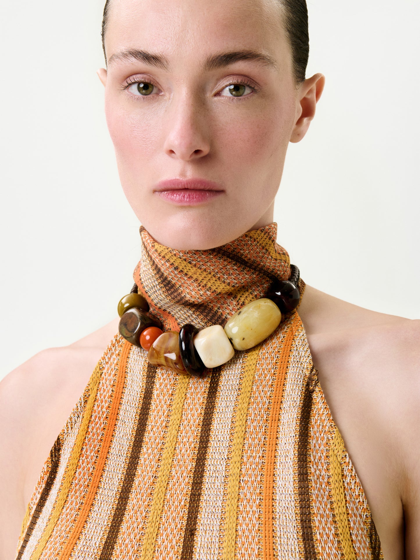 A woman with light skin and slicked-back hair wears the Mitzy Blouse—a sleeveless, high-neck top with orange, yellow, and brown stripes, a chunky beaded necklace, a true-to-size fit, and premium Italian fabric.