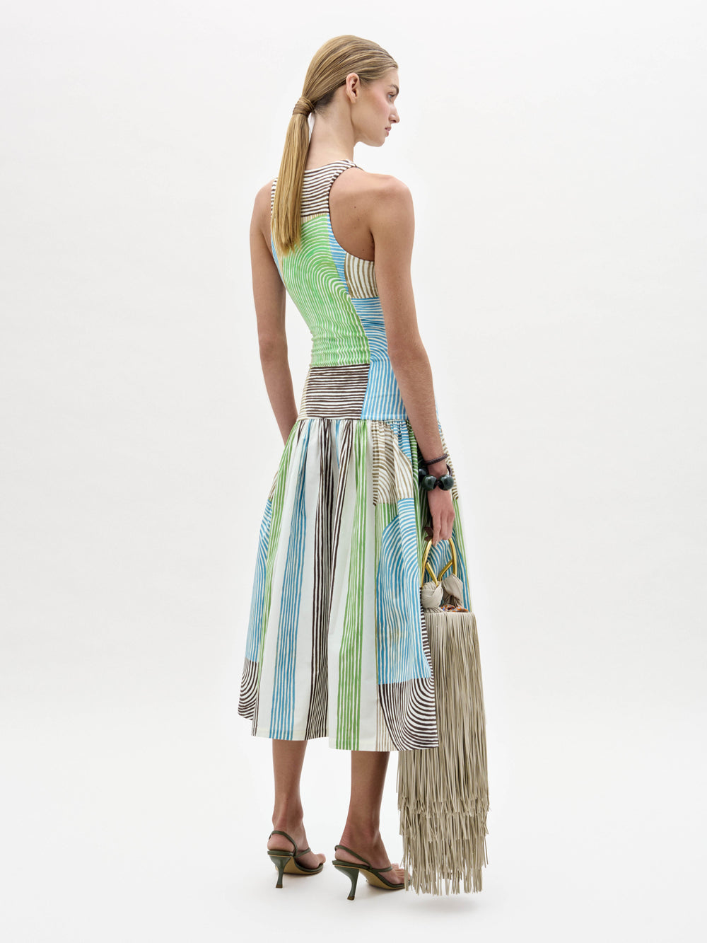 A woman models the Jamelia Dress, a sleeveless color-blocked midi with stripe patterns, styled with black heeled sandals and a fringed handbag, posing against a white background.